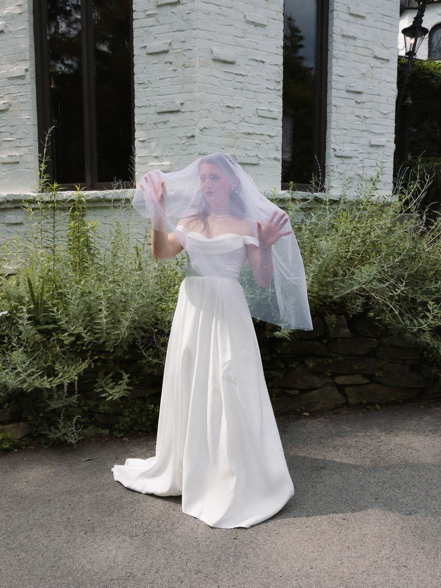 A bride in a flowing white wedding gown and veil poses outdoors against a white stone wall among garden greenery.