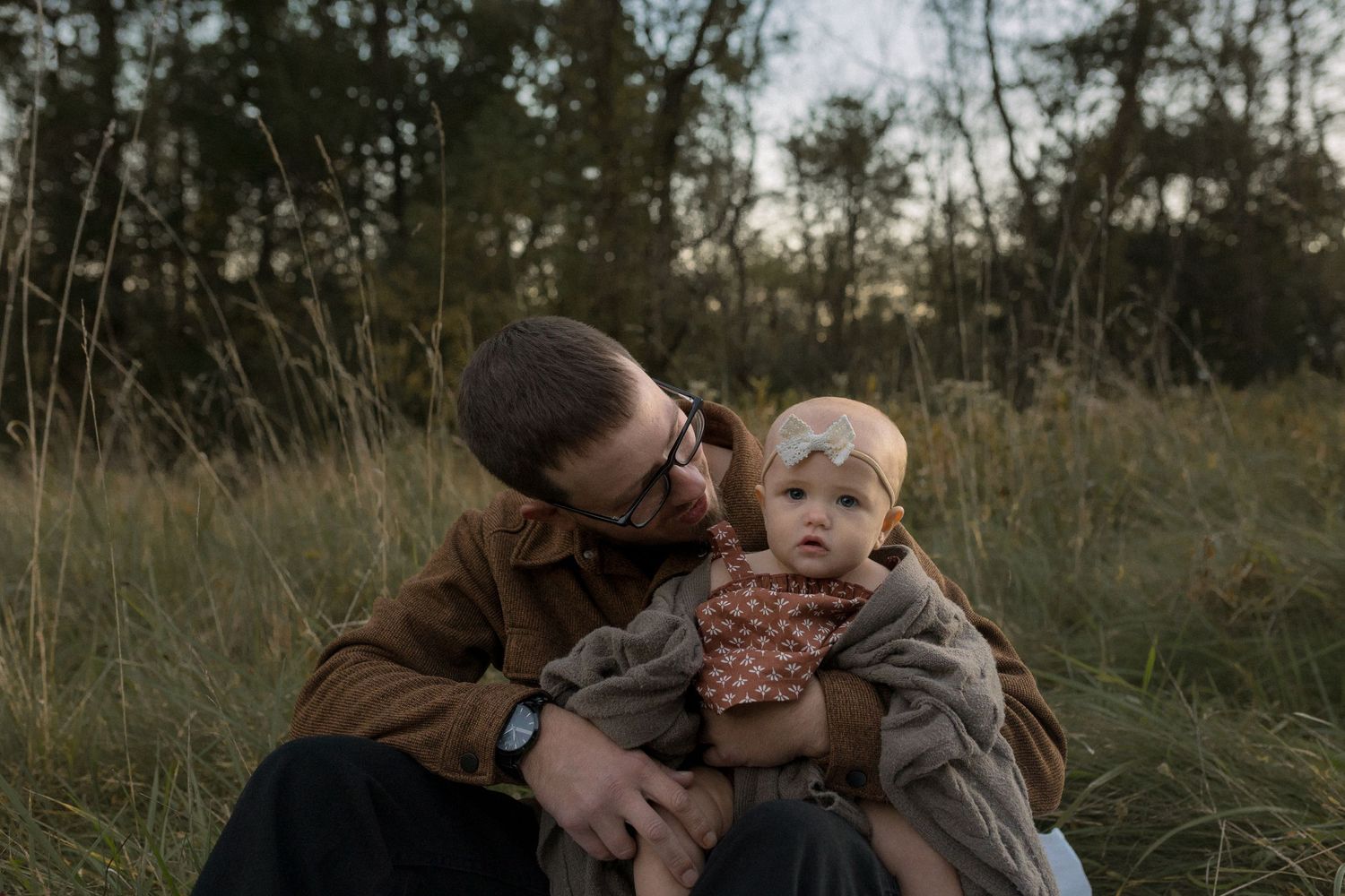 A tender moment shared sitting in tall grass at dusk with a baby wearing brown clothing.