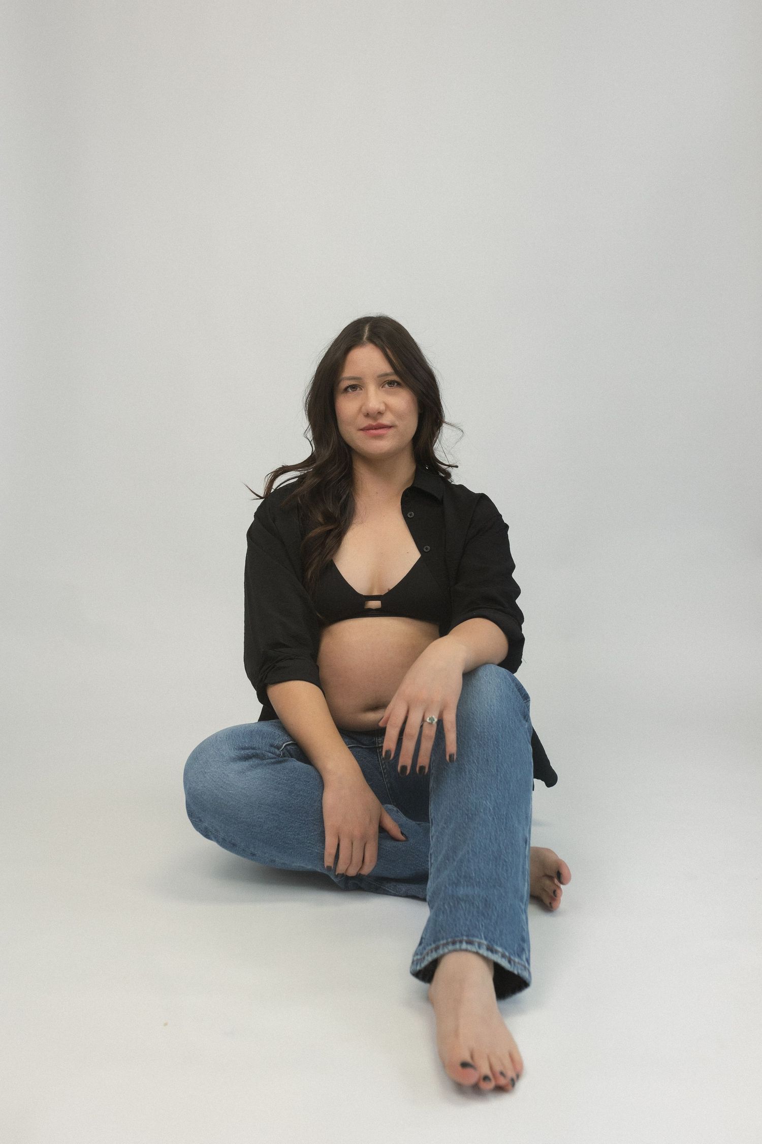 Woman in black top and jeans sitting cross-legged against white studio background.