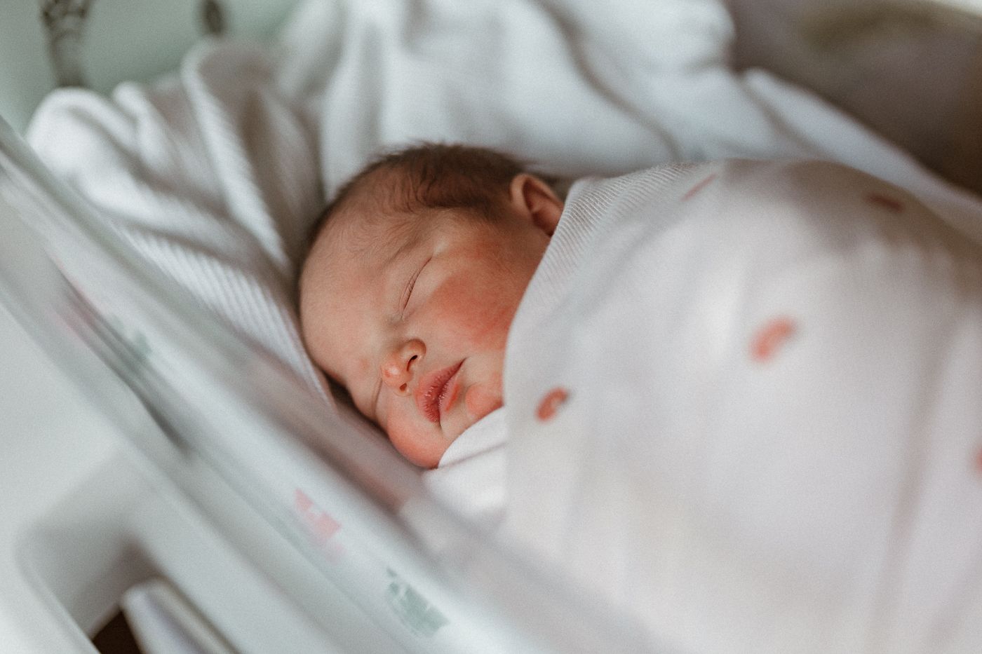 A sleeping newborn baby rests on white hospital linens with closed eyes and peaceful expression.