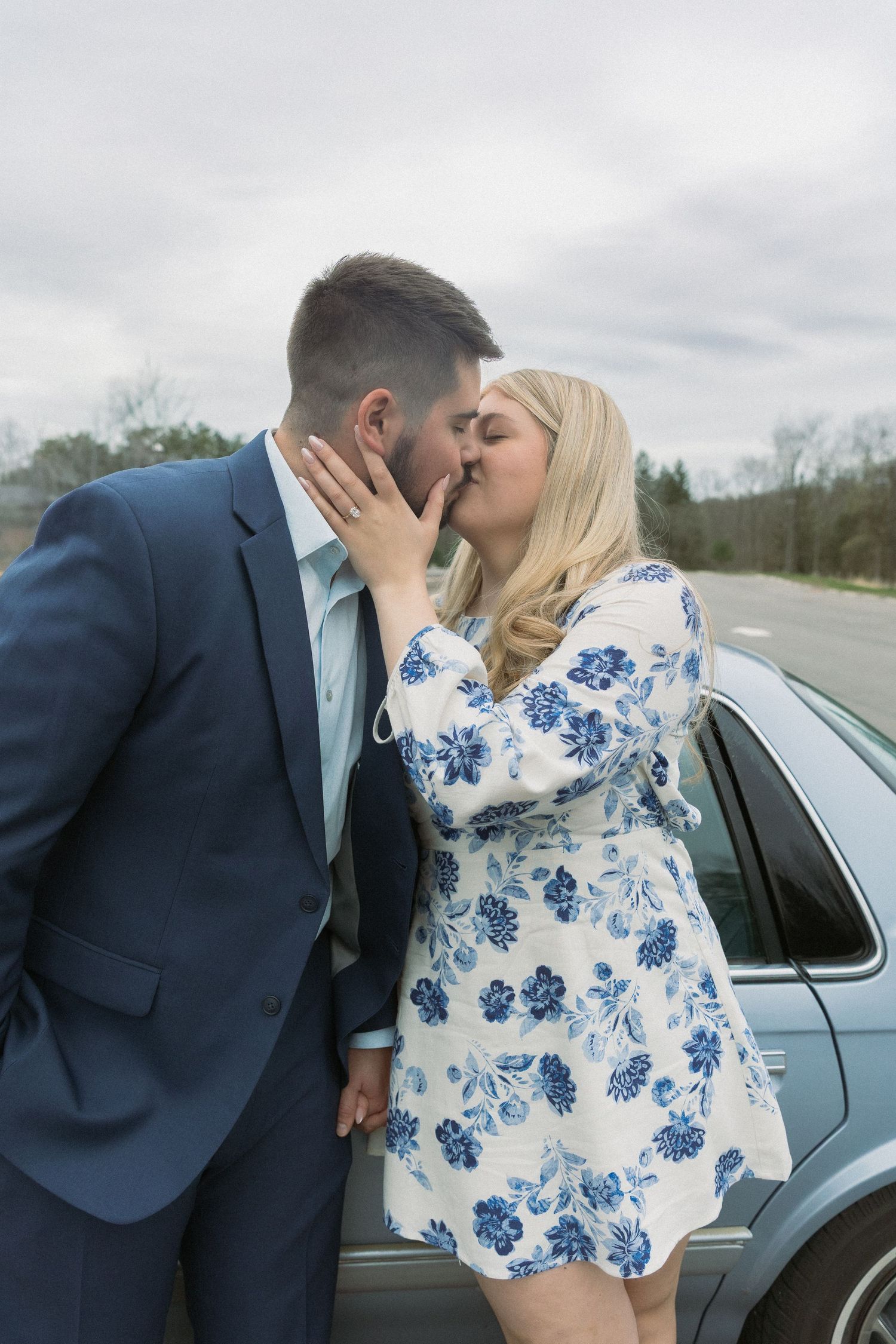 Romantic kiss during surprise proposal, couple styled in blue and white beside a vintage car, candid engagement photography.
