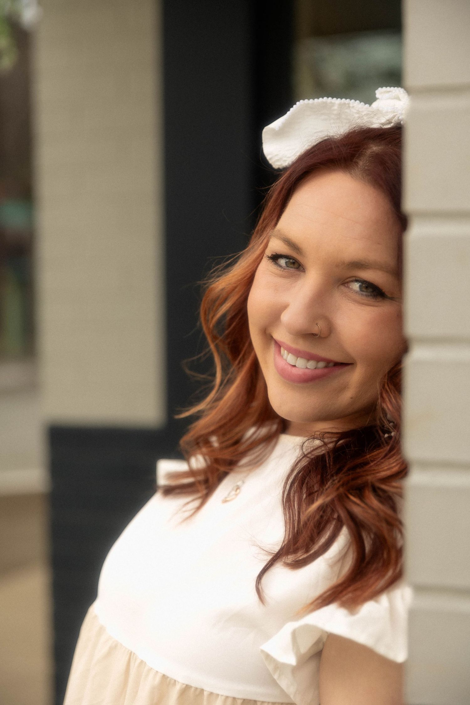 Woman in white vintage-style dress and hair bow poses elegantly against building exterior.