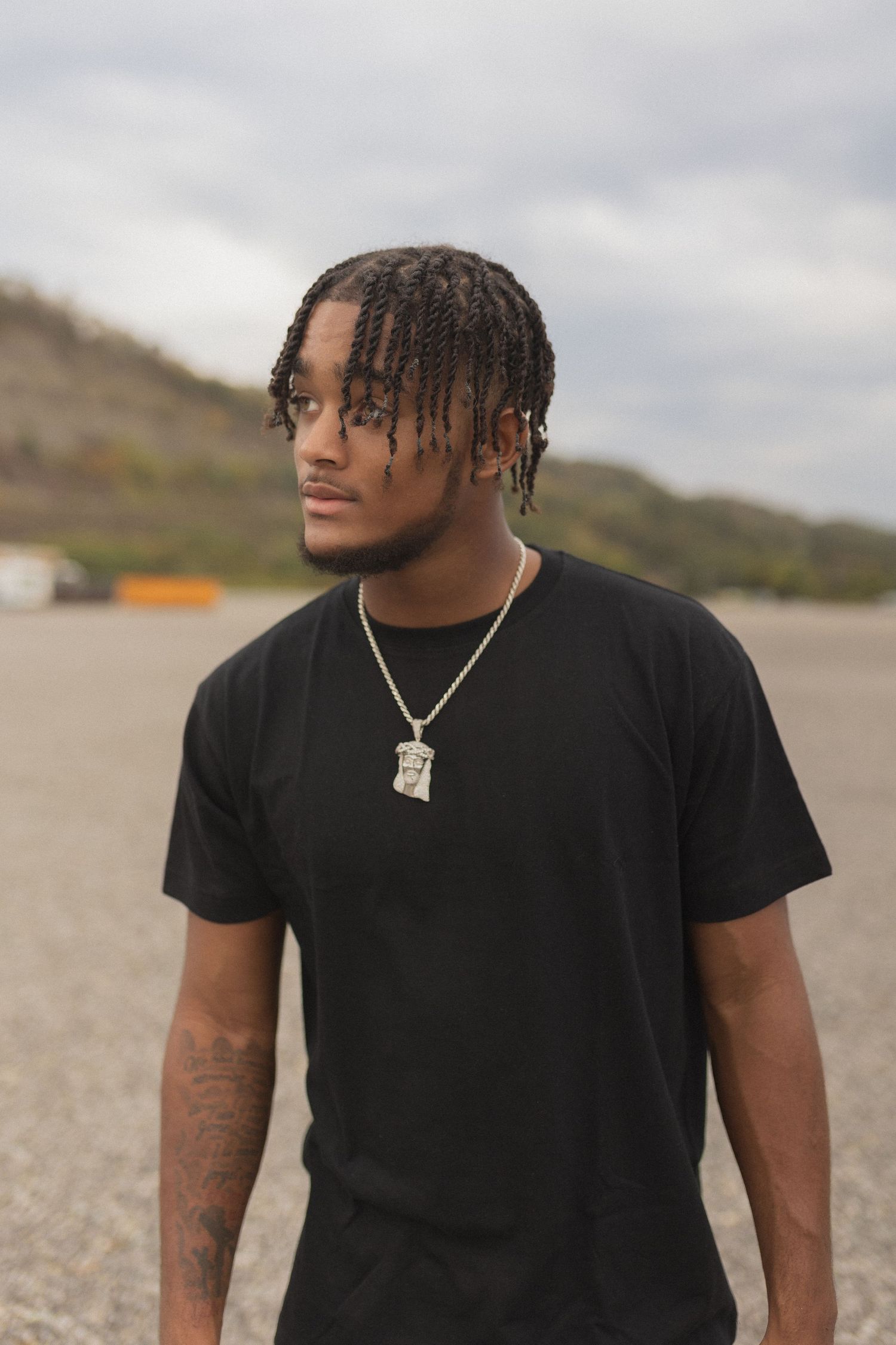 Portrait of person in black t-shirt with braided hair and necklace outdoors.