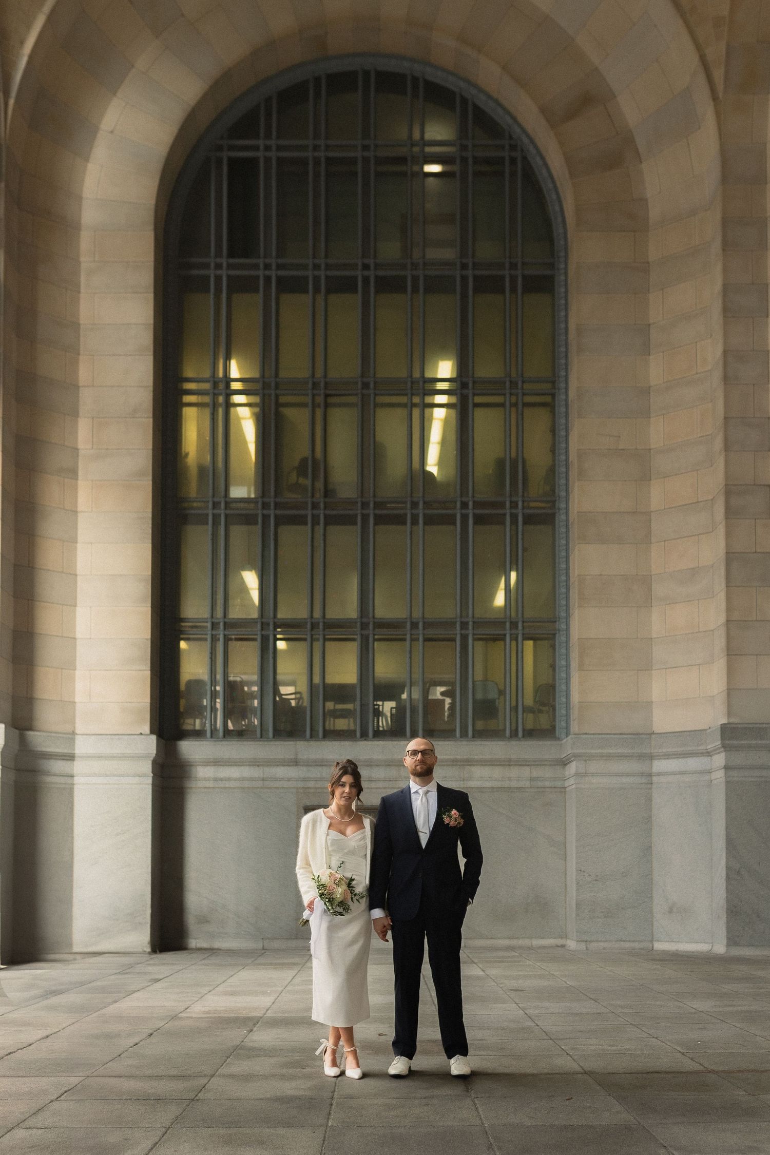 A newlywed couple standing under a grand stone archway in formal wedding attire.