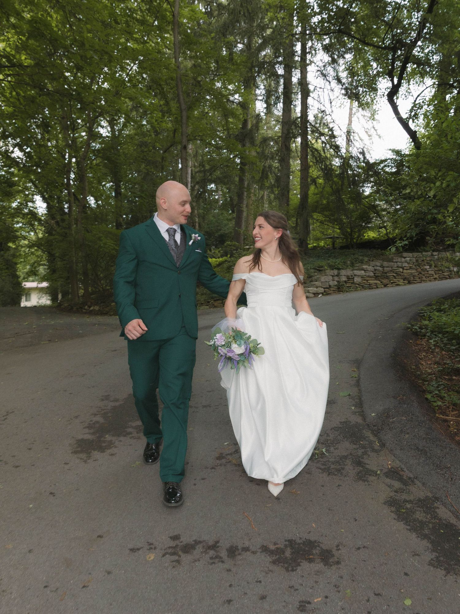A couple walking together on a winding path with trees and stone wall, wearing a green suit and white wedding dress.
