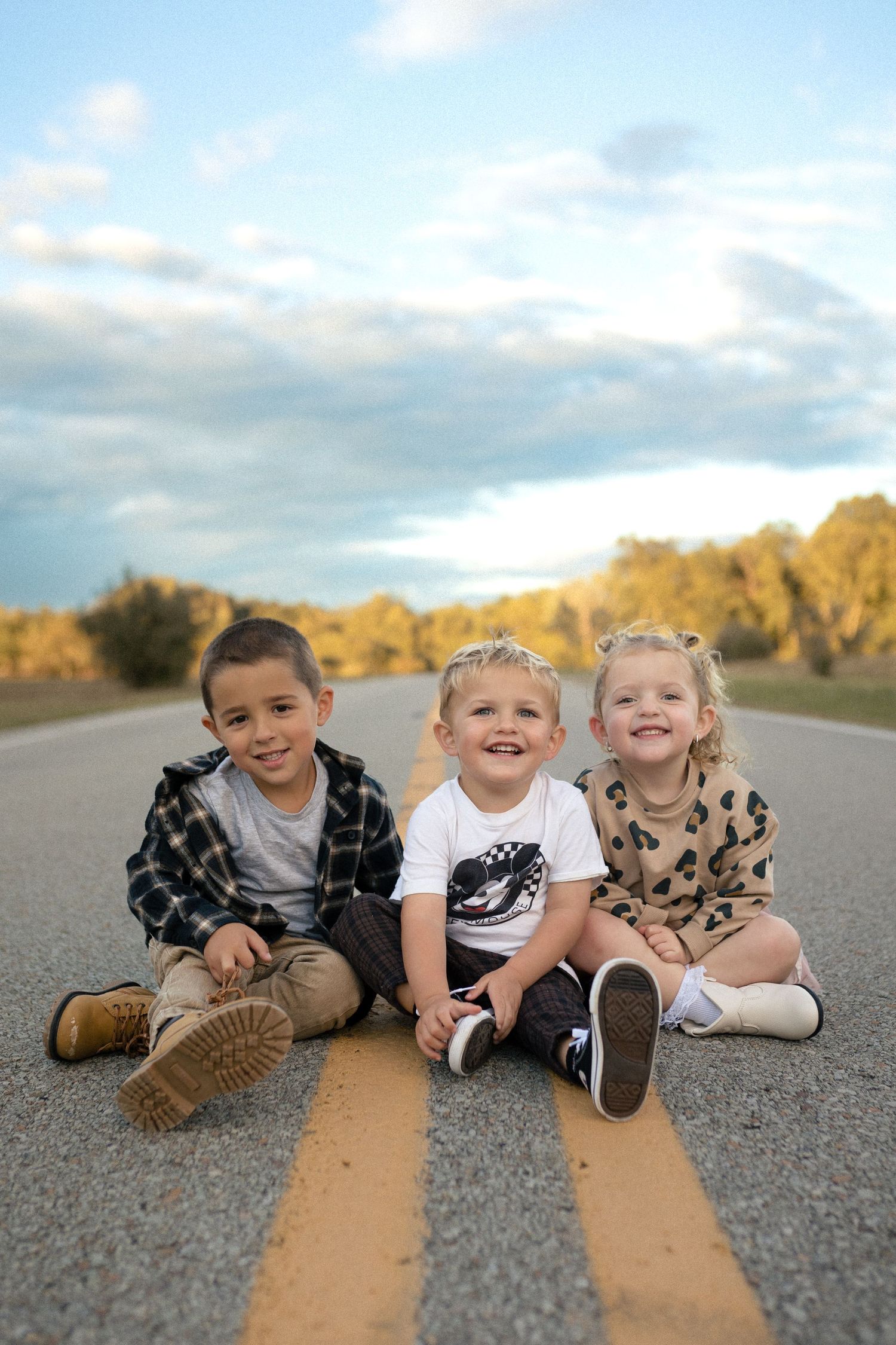 Three children sitting together on yellow road lines during sunset.