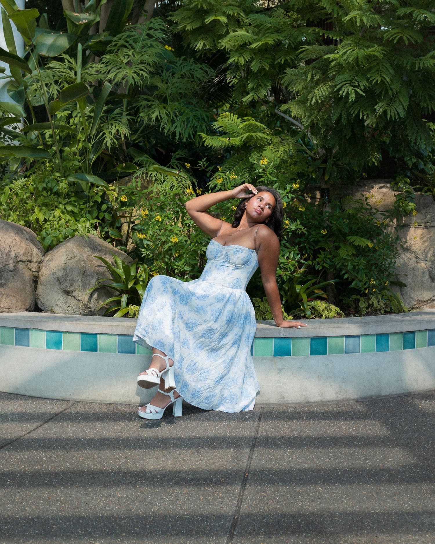 A person in a blue dress lays gracefully along a curved pool edge surrounded by lush green foliage.