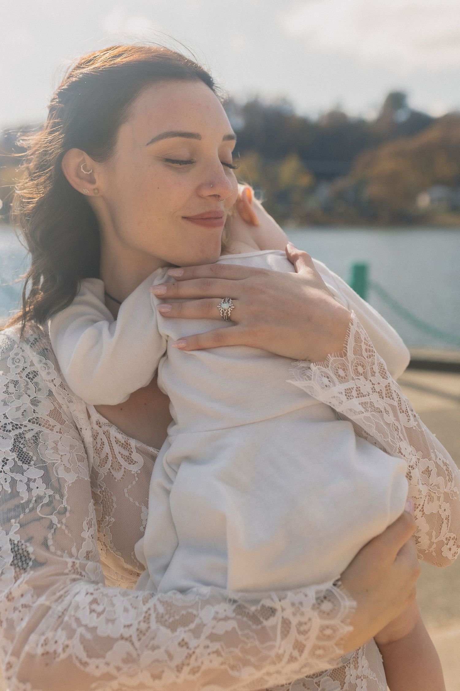 A tender moment between a bride in lace holding a baby against a waterfront background.