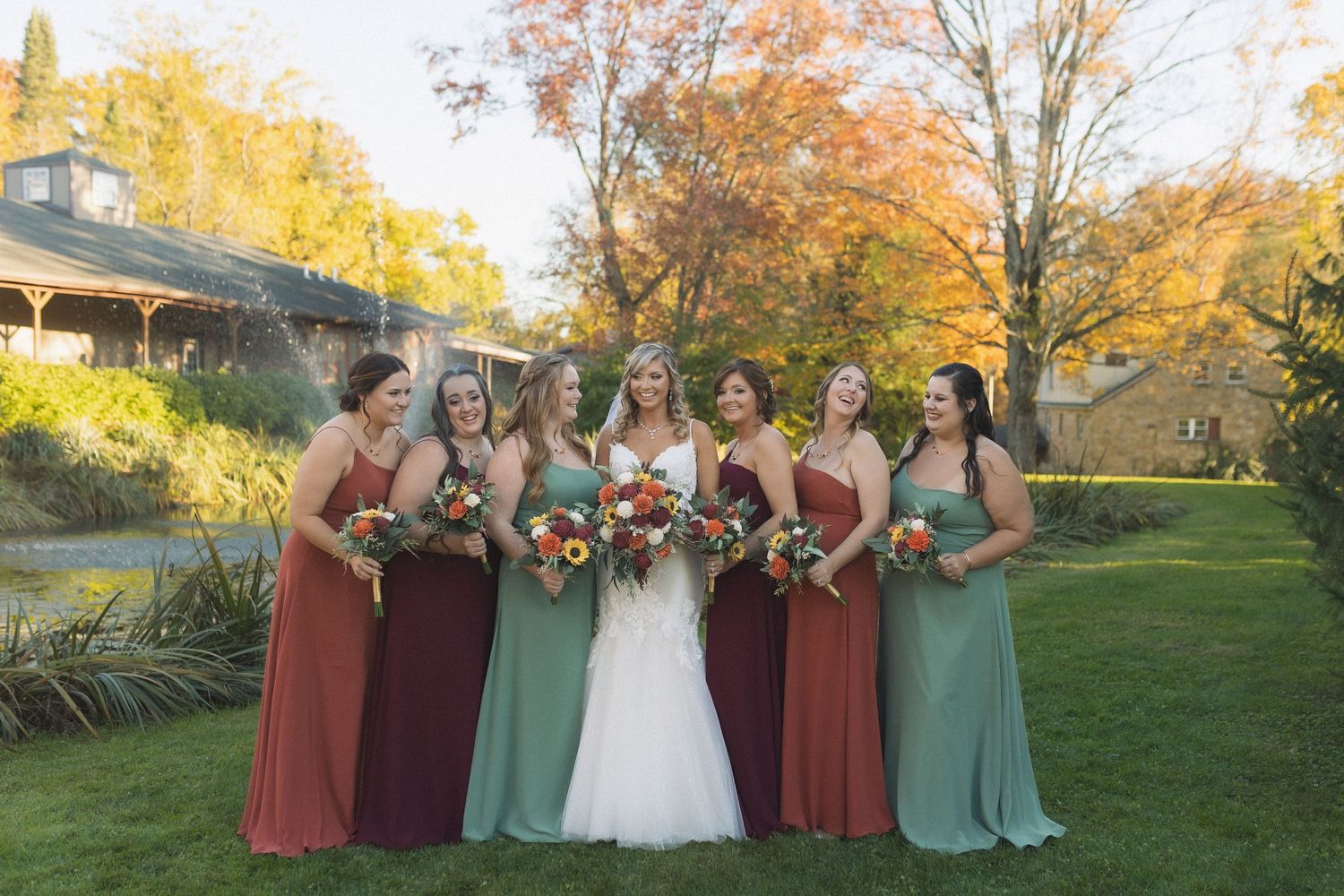 Wedding party in burgundy and sage green dresses pose together on a lawn with autumn foliage in the background.