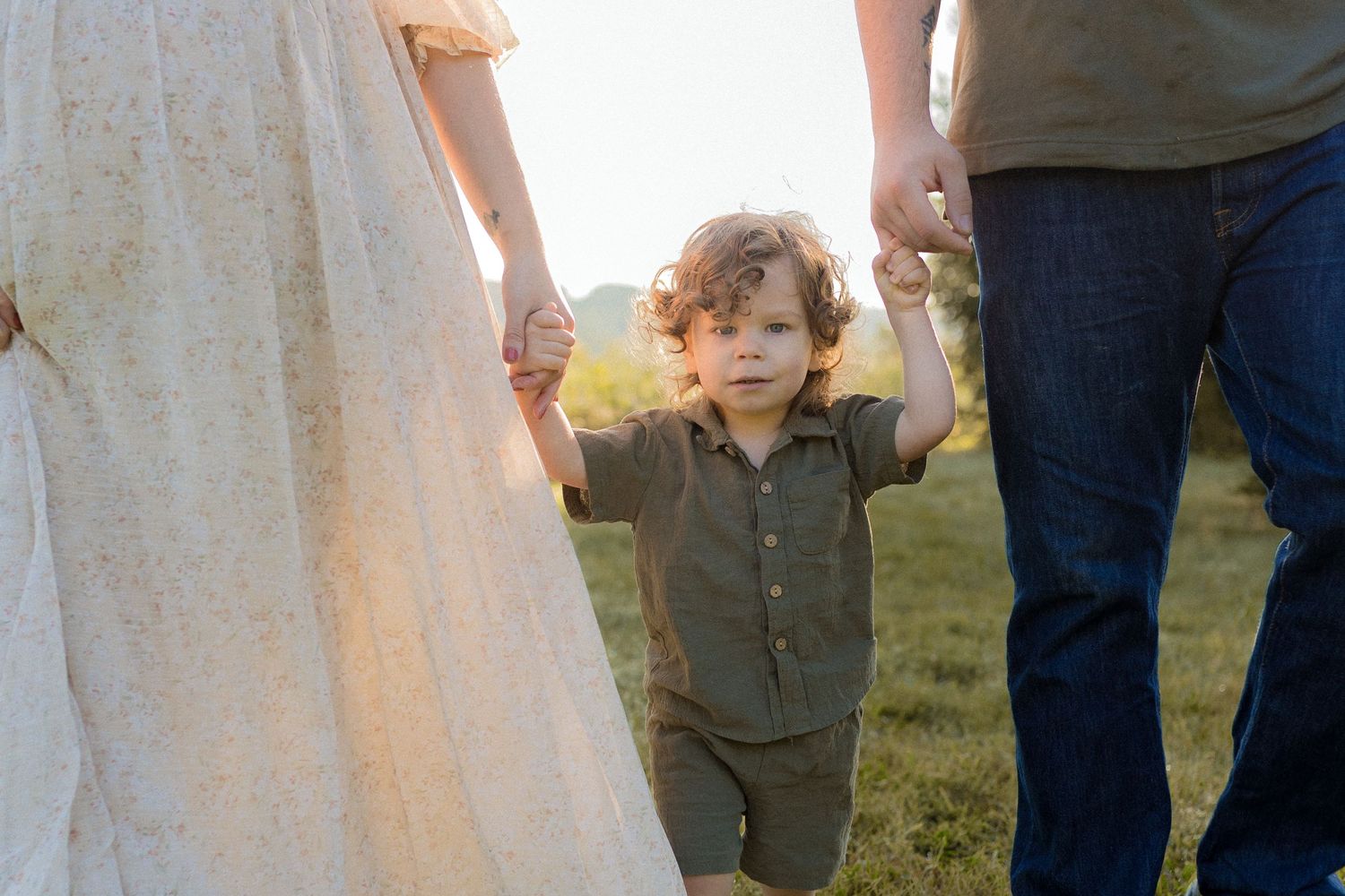 Small child walking while holding hands with parents in a grassy field at sunset.