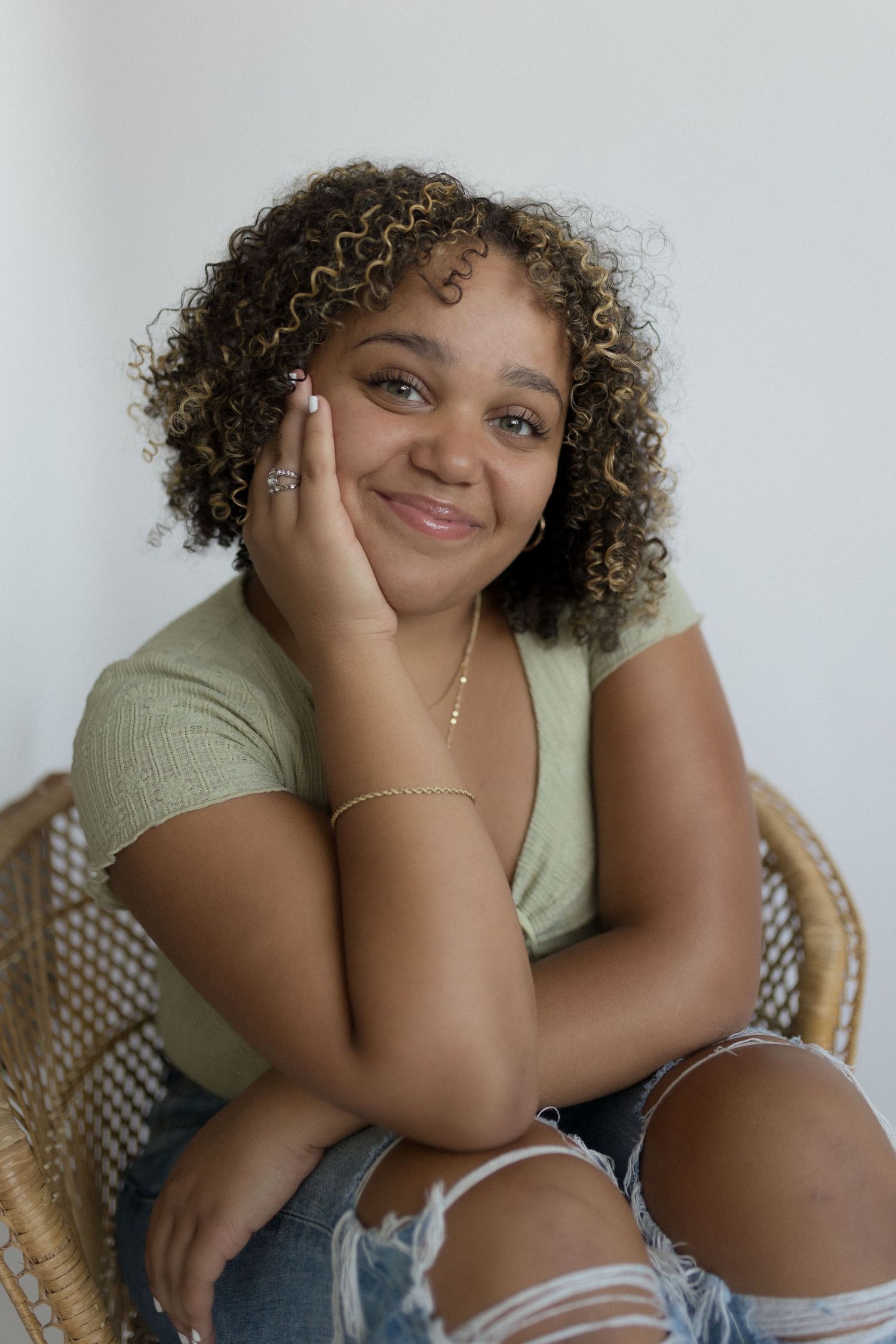 Person in green top and ripped jeans sitting in wicker chair against white background.