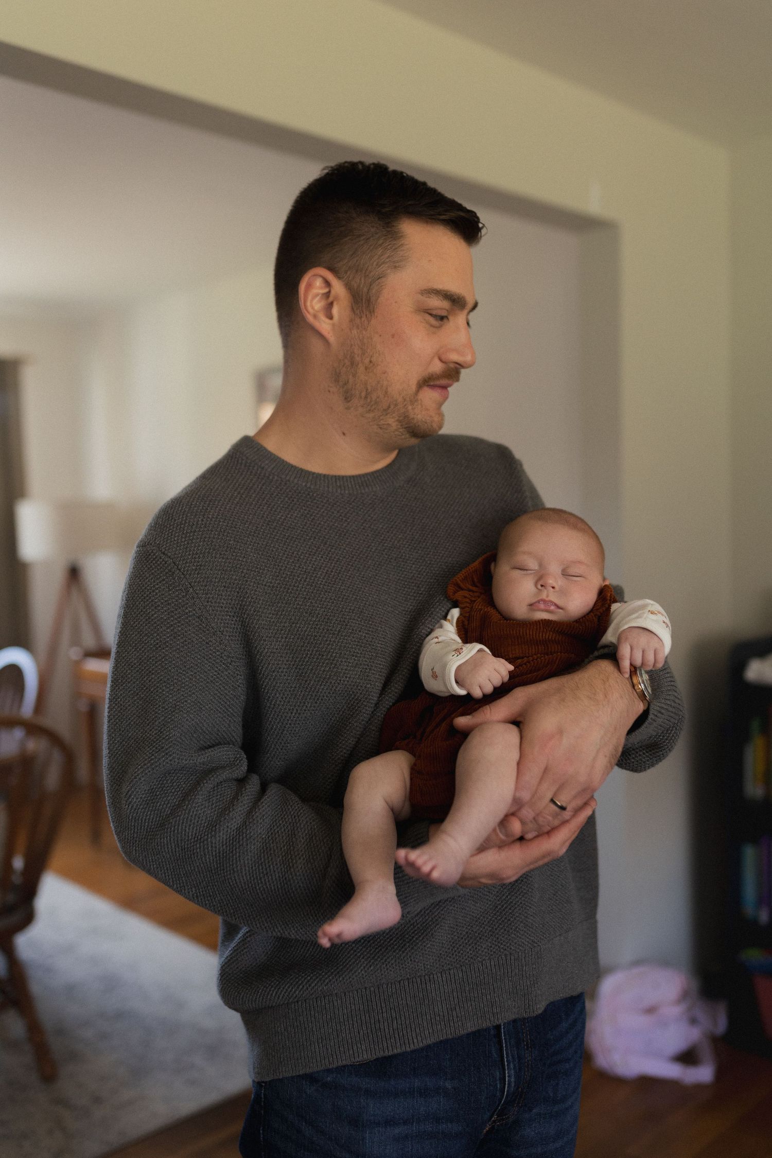 A sleeping infant in brown overalls is held gently in a cozy home setting.