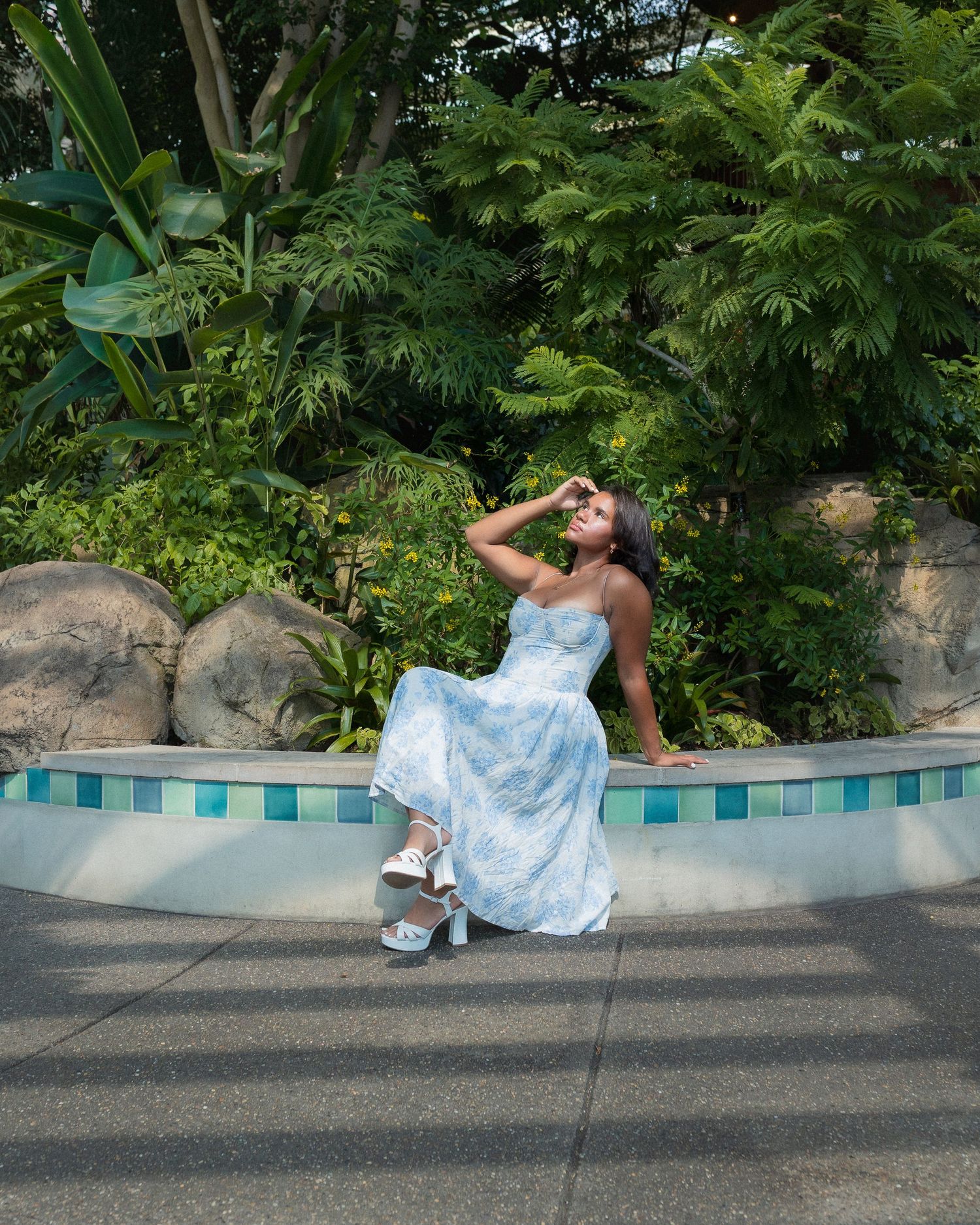 Someone in a light blue floral dress sits on a curved tiled wall surrounded by lush tropical greenery and plants.