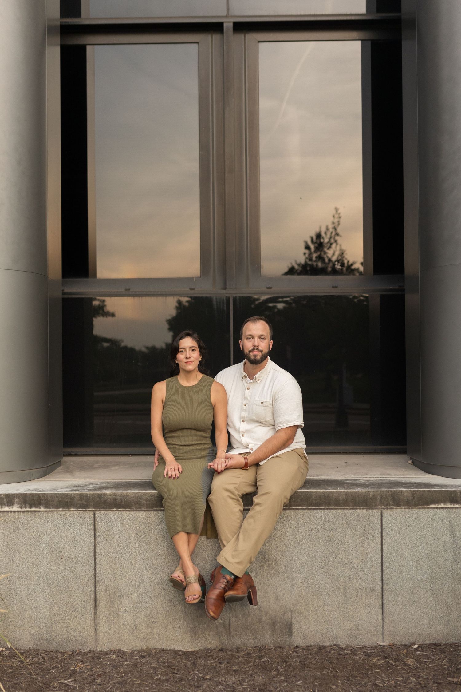 Modern engagement session with clean lines and soft film tones, couple seated casually in front of a mirrored building.