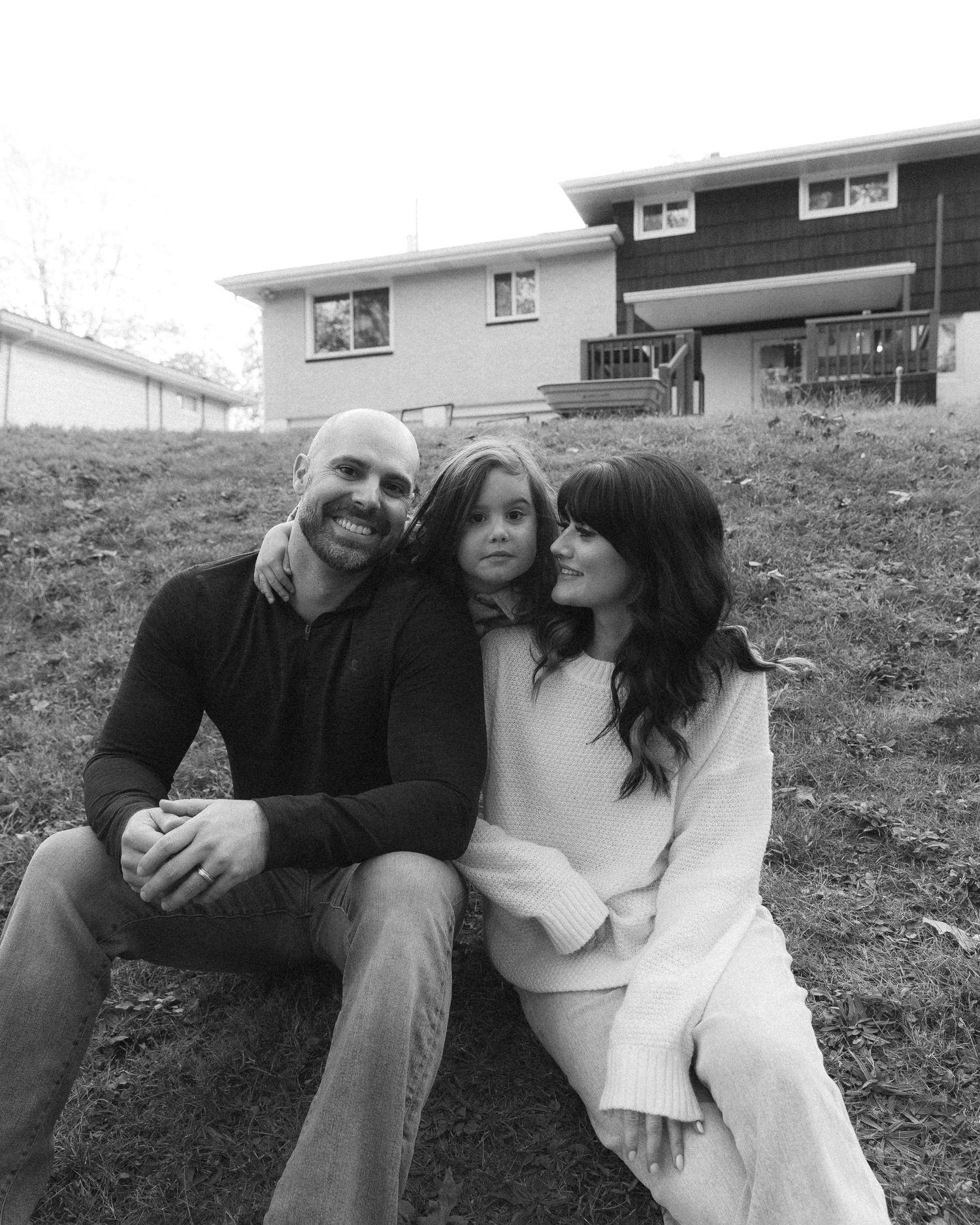 A black and white photo of three people sitting together on a grassy hill in front of a house.