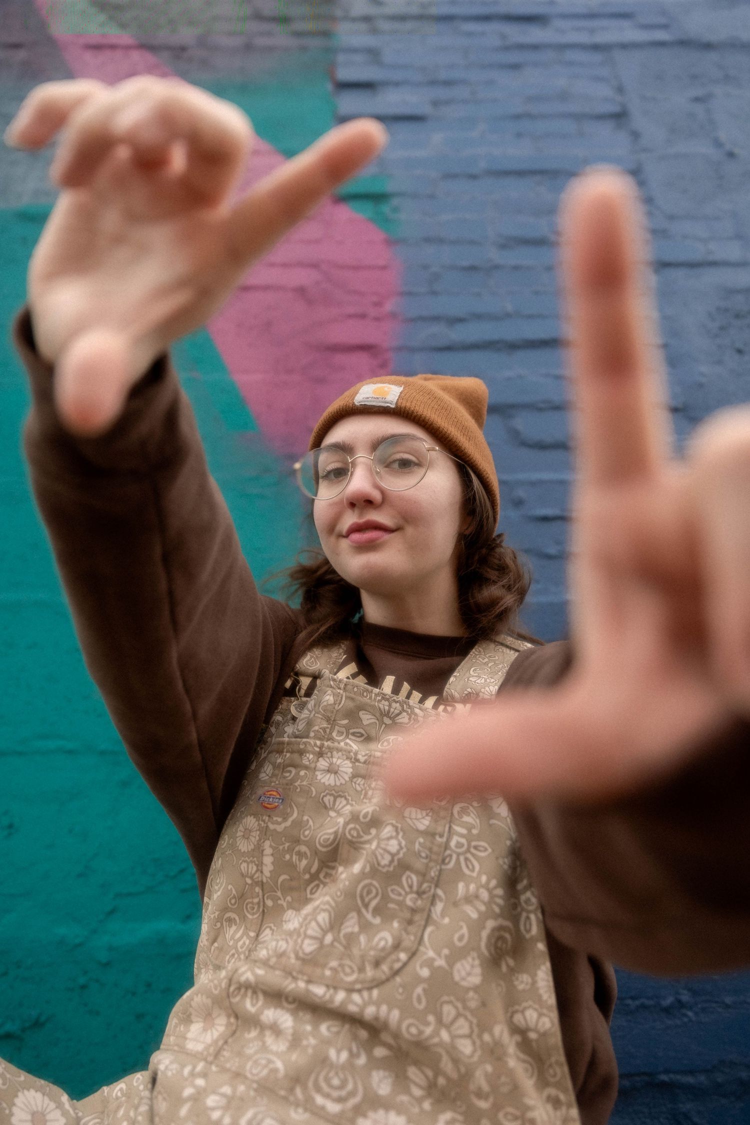 Person in brown beanie and overalls makes framing gesture with hands against colorful wall.