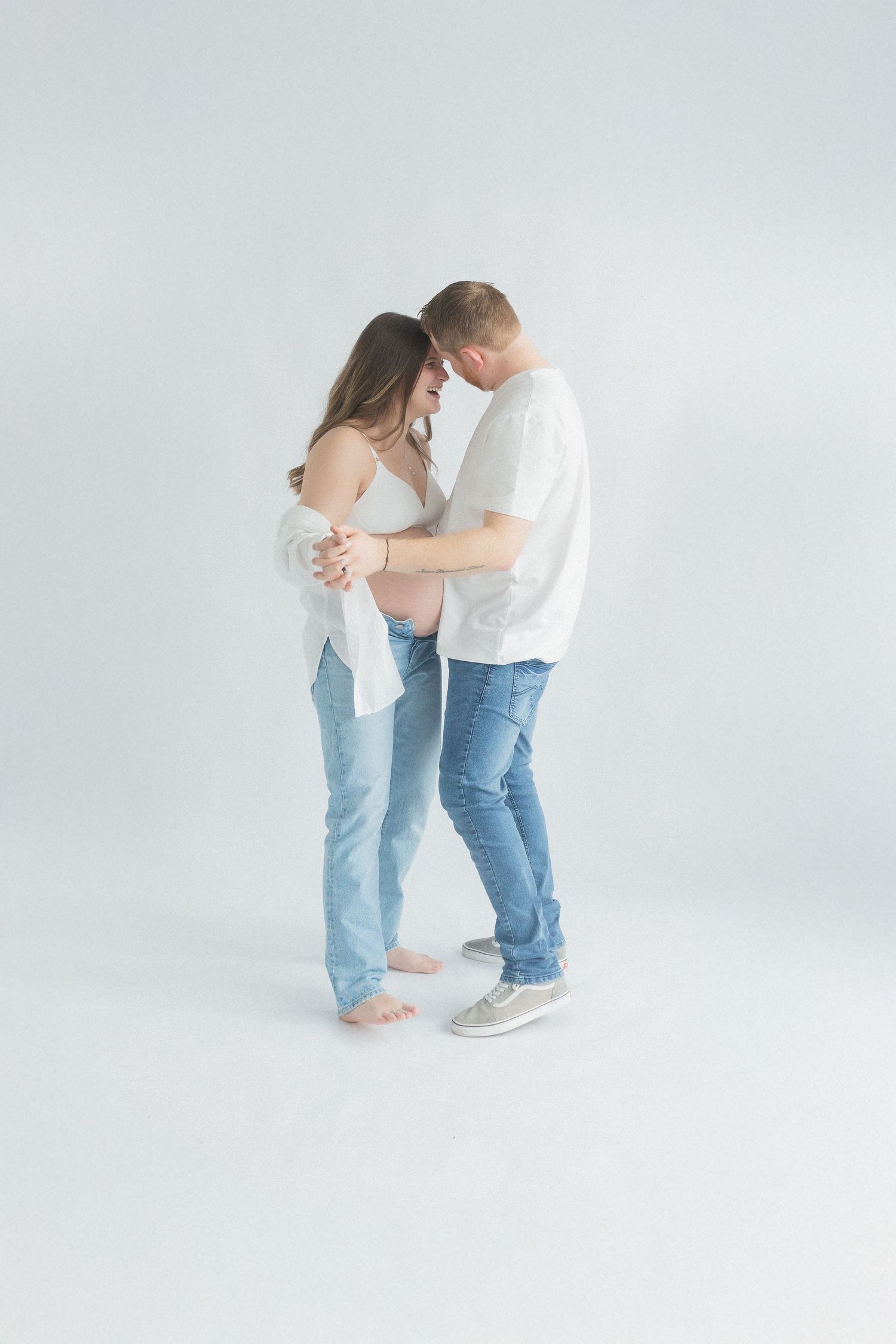 Couple shares an intimate dance-like moment in matching jeans and white tops against plain background.