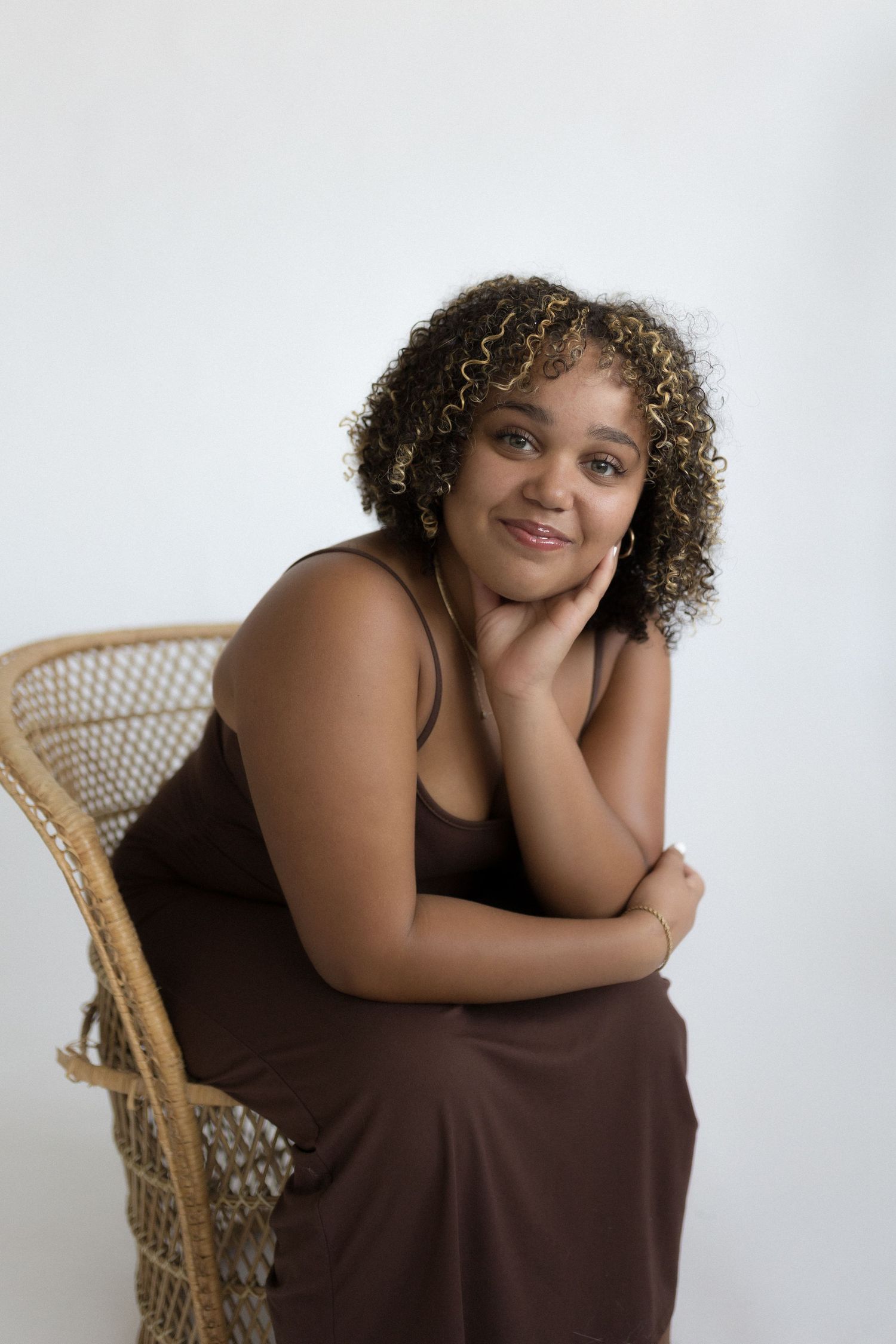 Person sitting in woven chair wearing brown dress during studio photoshoot.