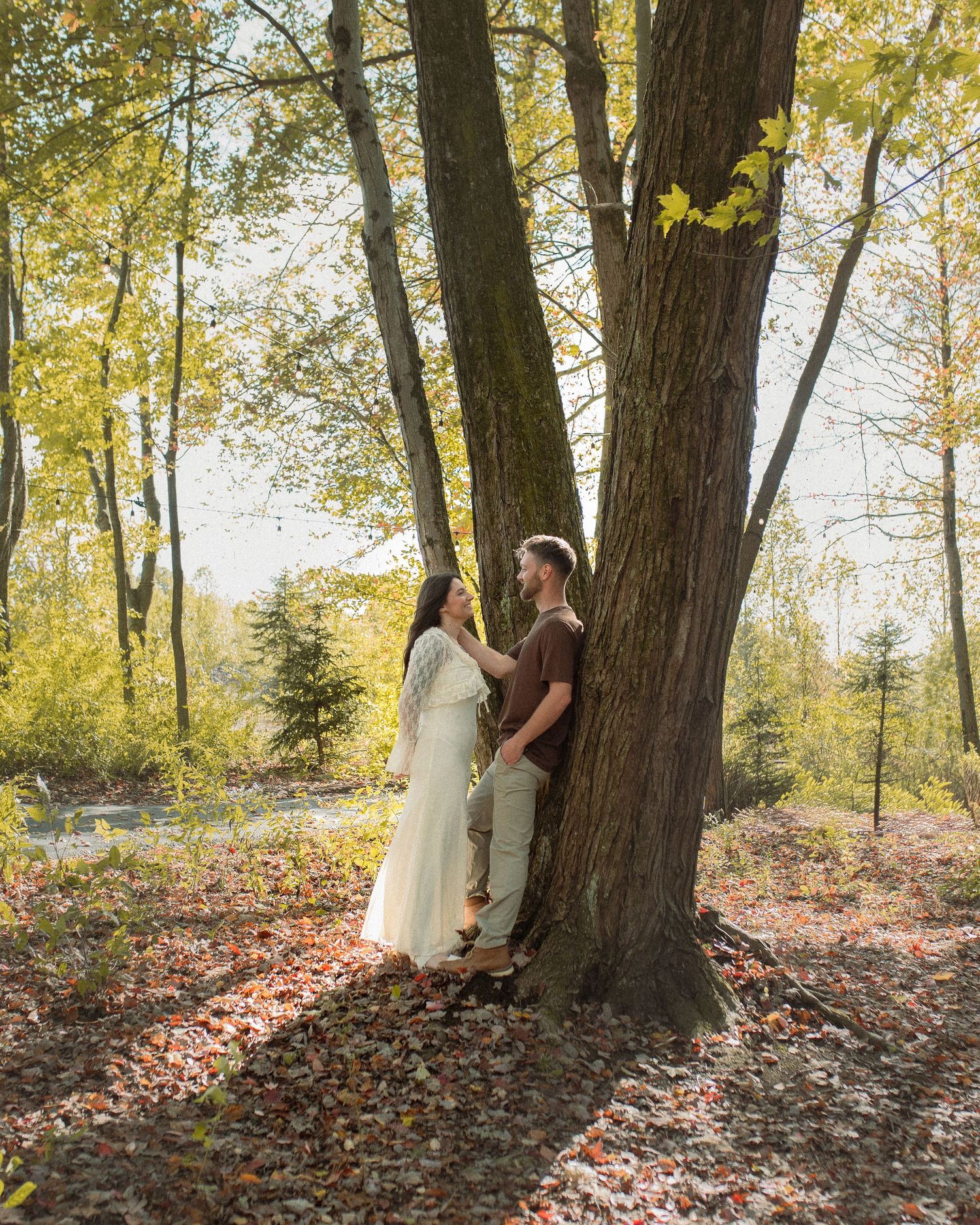 A couple shares a tender moment against a tree trunk in an autumn forest setting with fallen leaves on the ground.