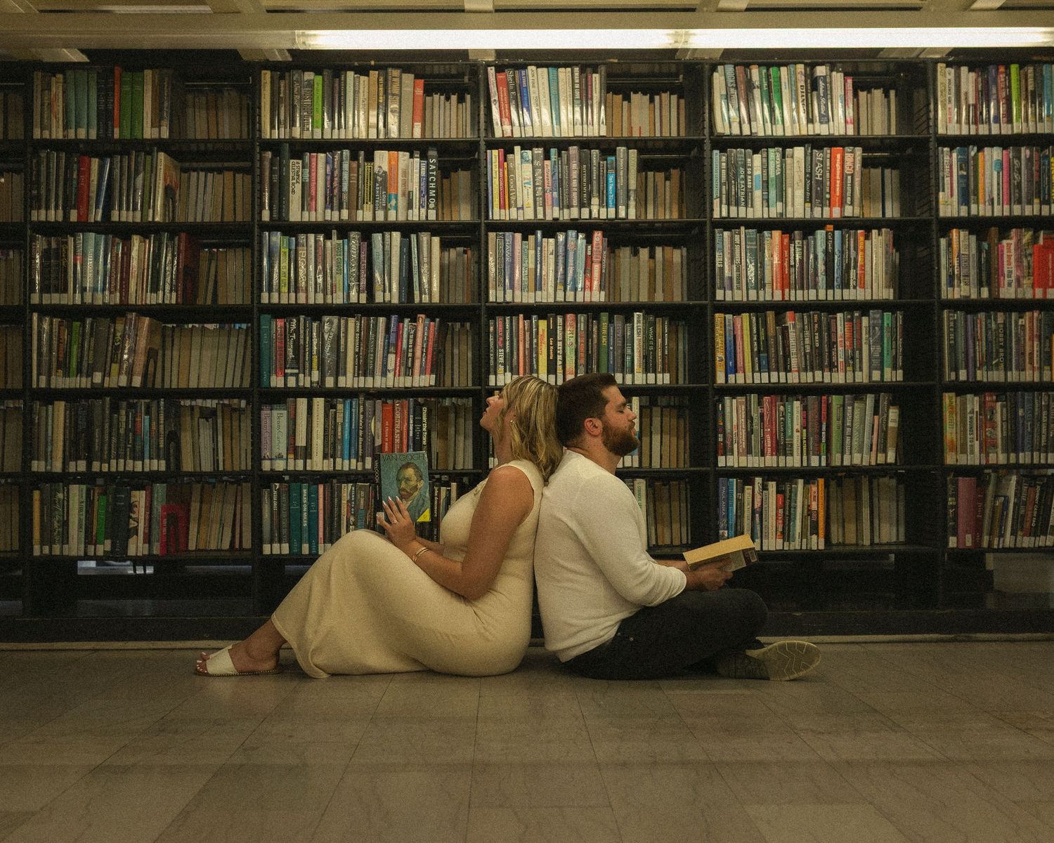 A series of bookshelves in a library with someone reclining on the floor between the stacks.