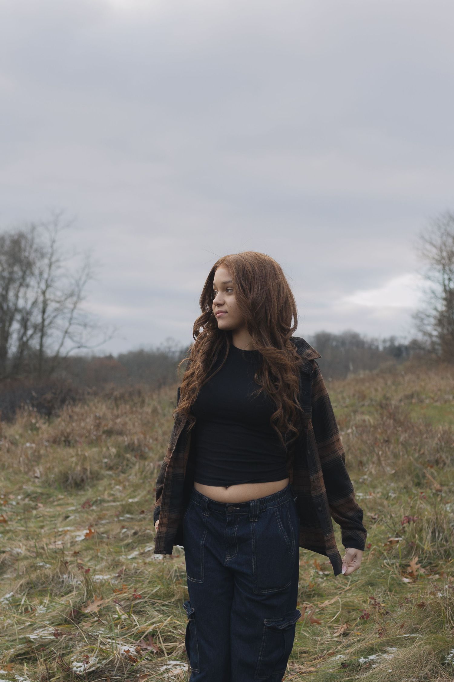 Person in black crop top and plaid jacket stands in autumn field under cloudy sky.