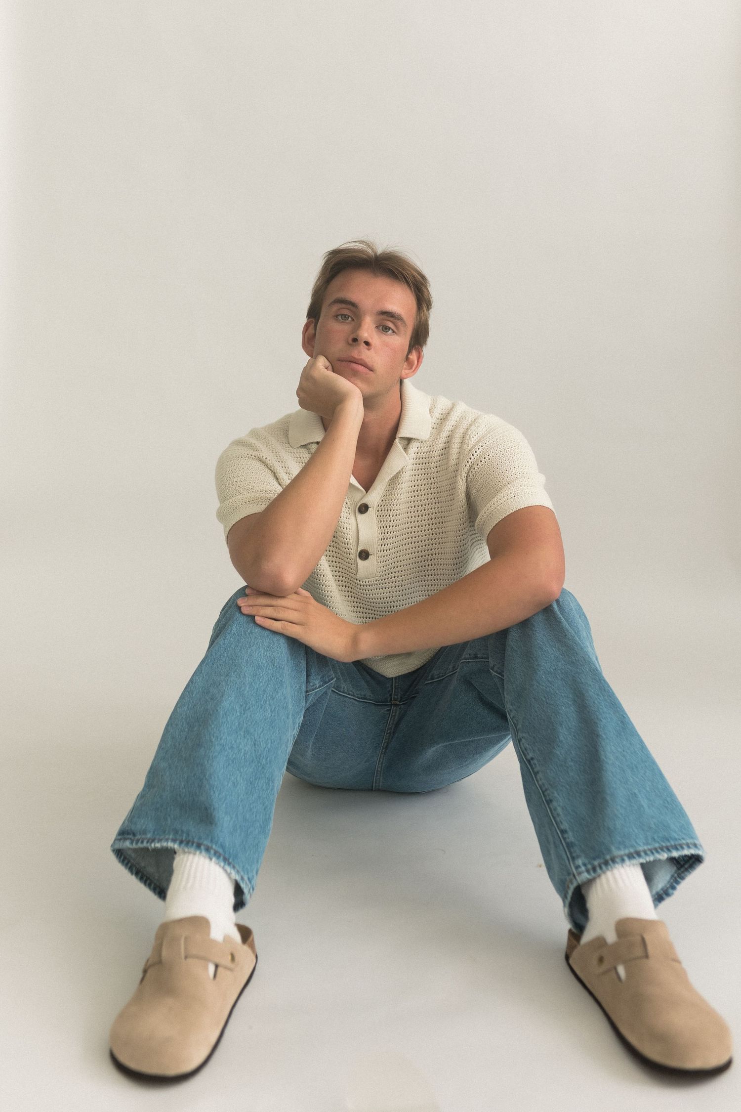 Model sits on floor wearing cream knit polo and relaxed jeans against white background.