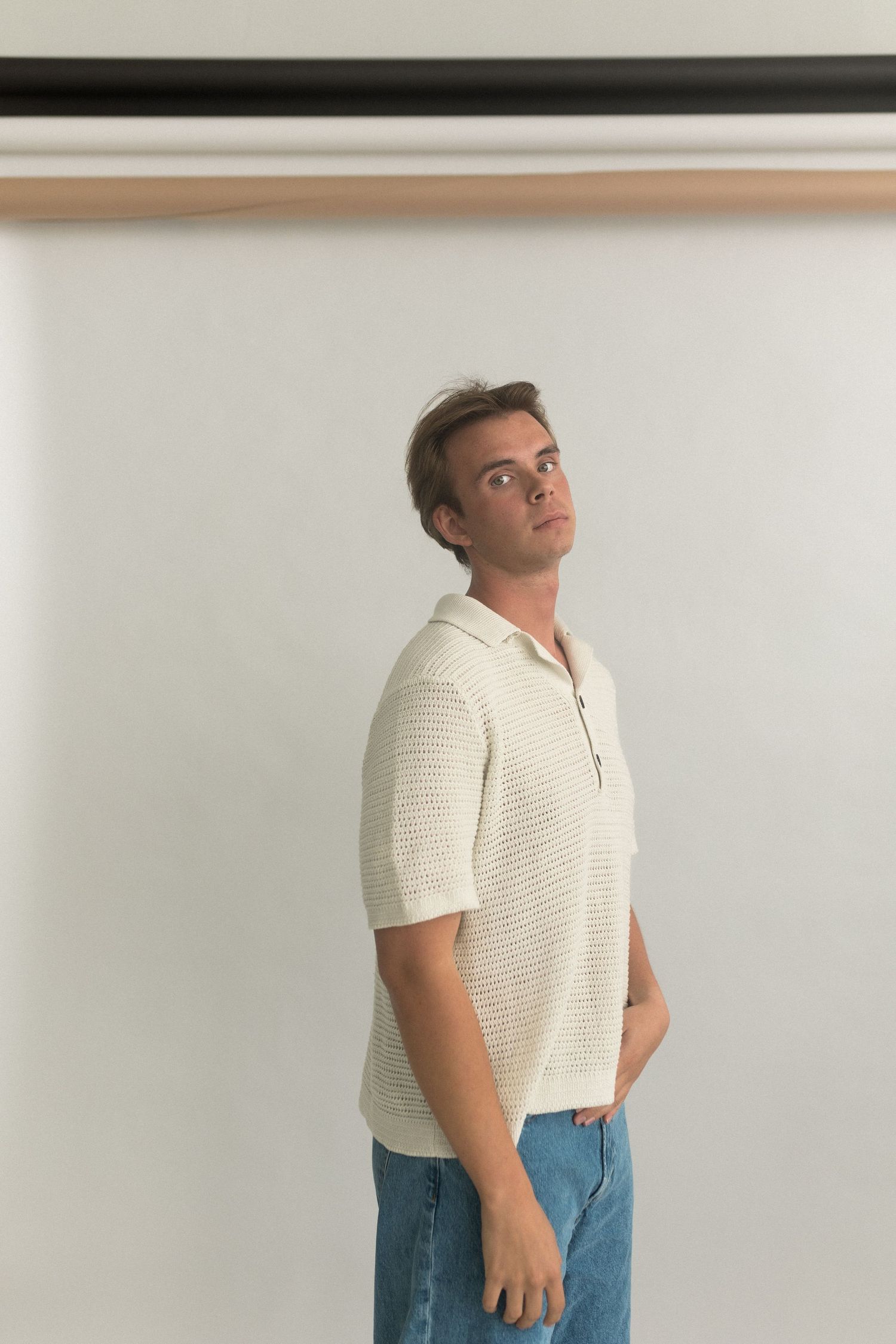 Person poses in cream knit polo shirt and blue jeans against white studio backdrop.
