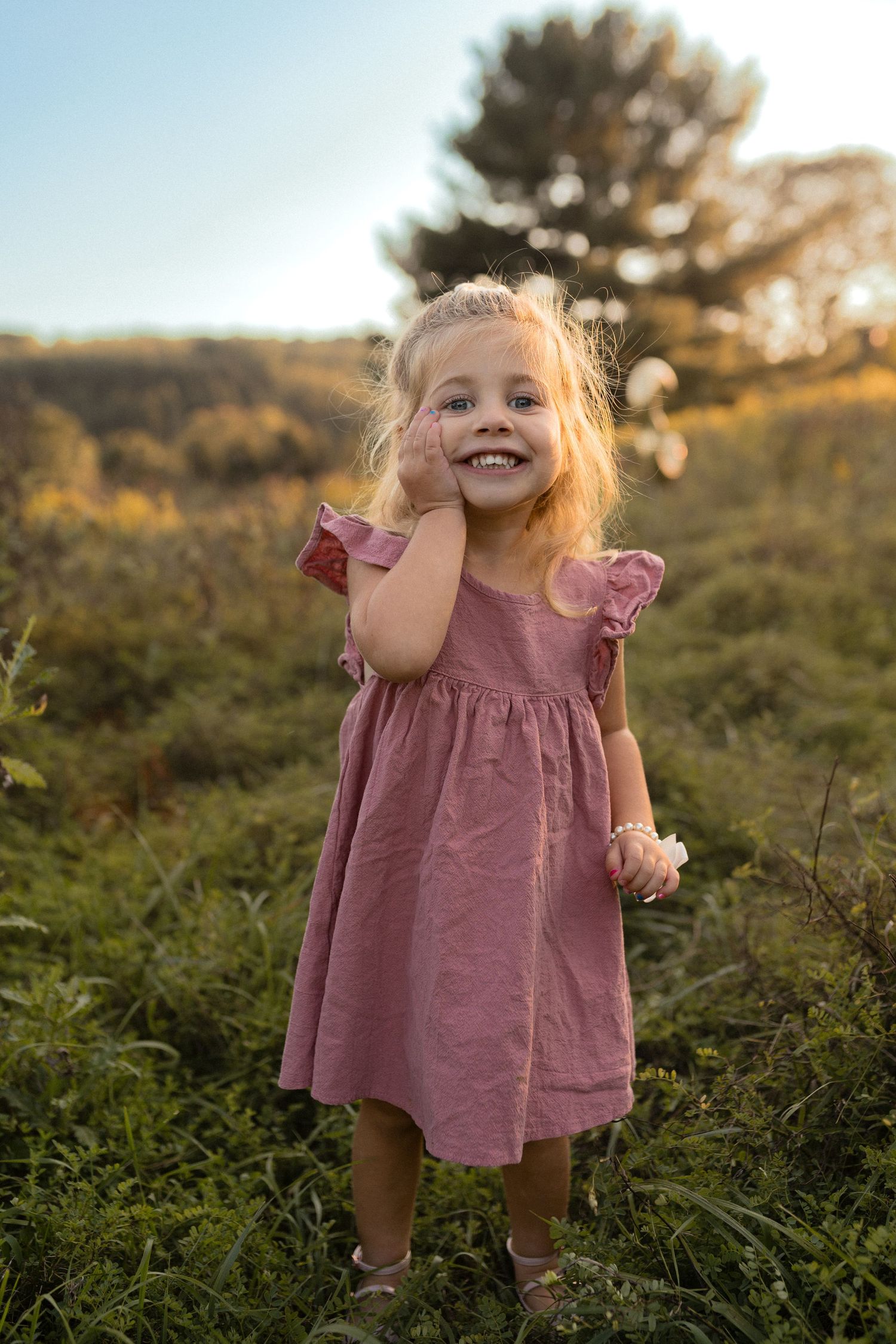 Child wearing dusty pink dress stands joyfully in sun-dappled field during golden hour.