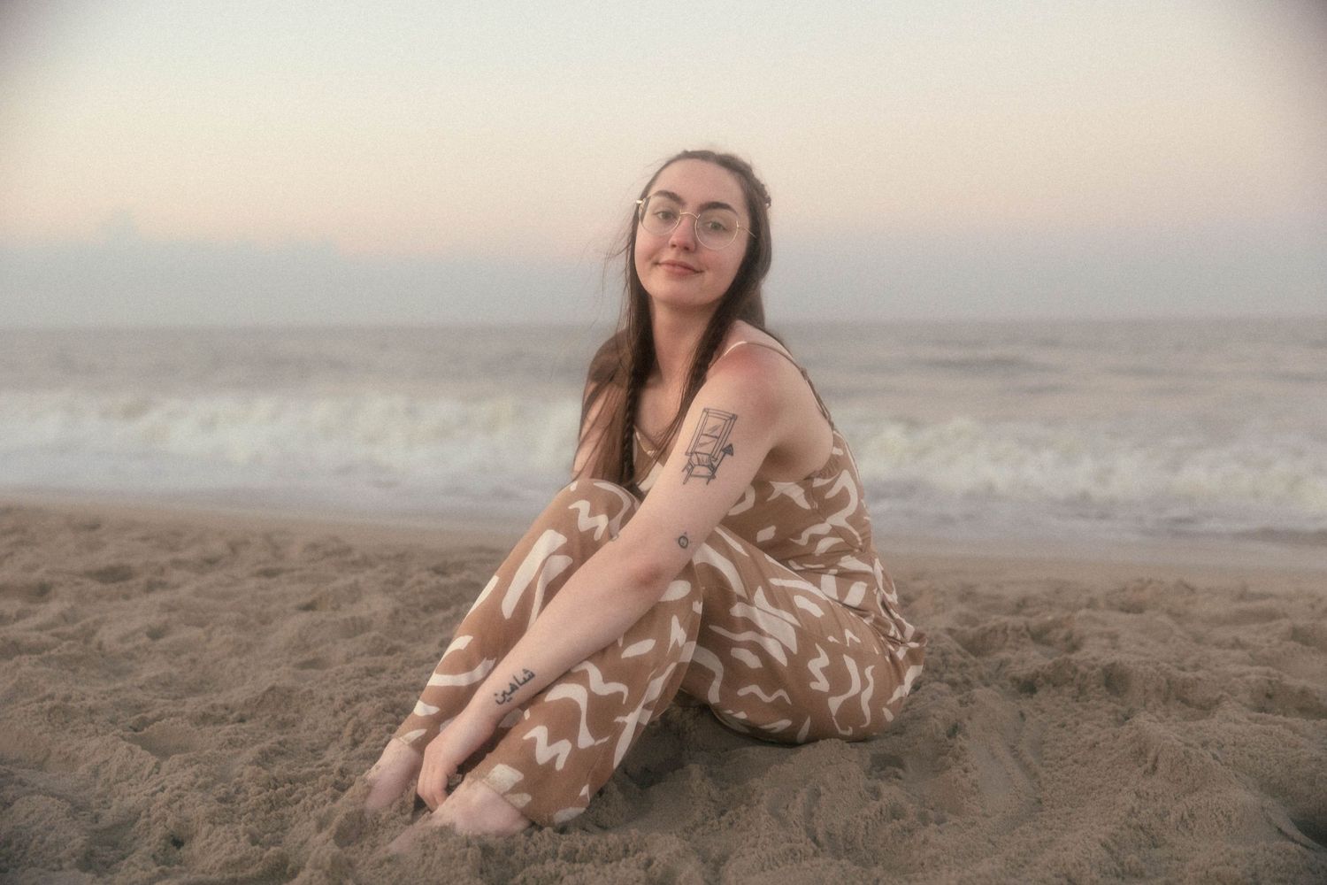 A person sits on sandy beach wearing a flowing tan dress with abstract white pattern.
