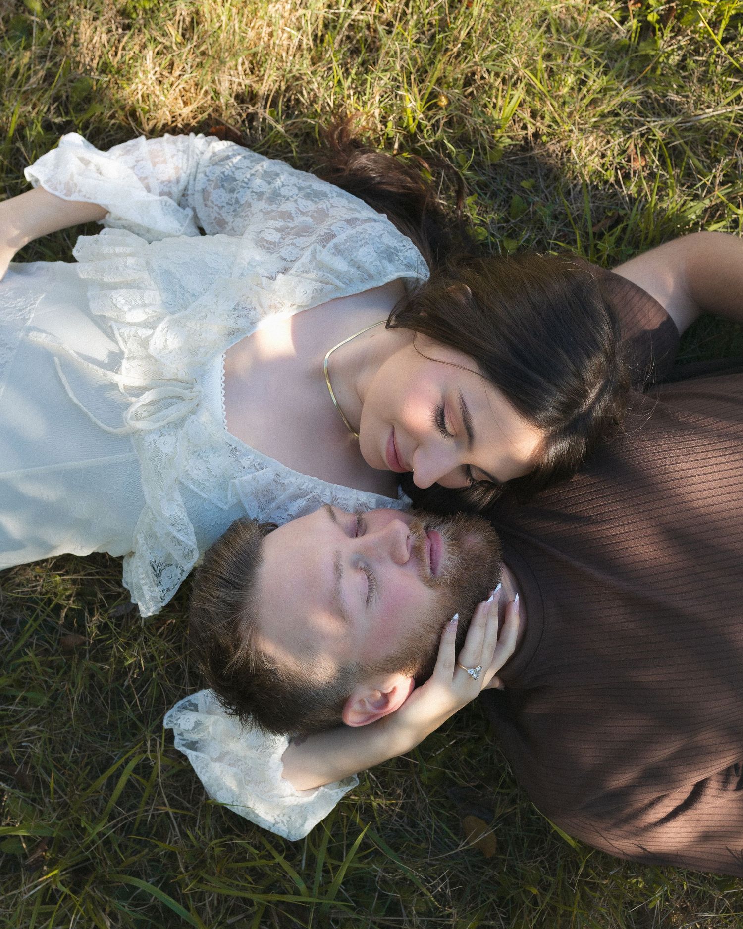 A person in white lace dress lies peacefully in grass with dappled sunlight streaming through tree branches above.