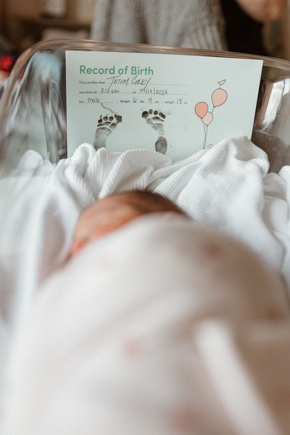 A birth record document with tiny footprints and balloon drawings rests above a swaddled newborn.
