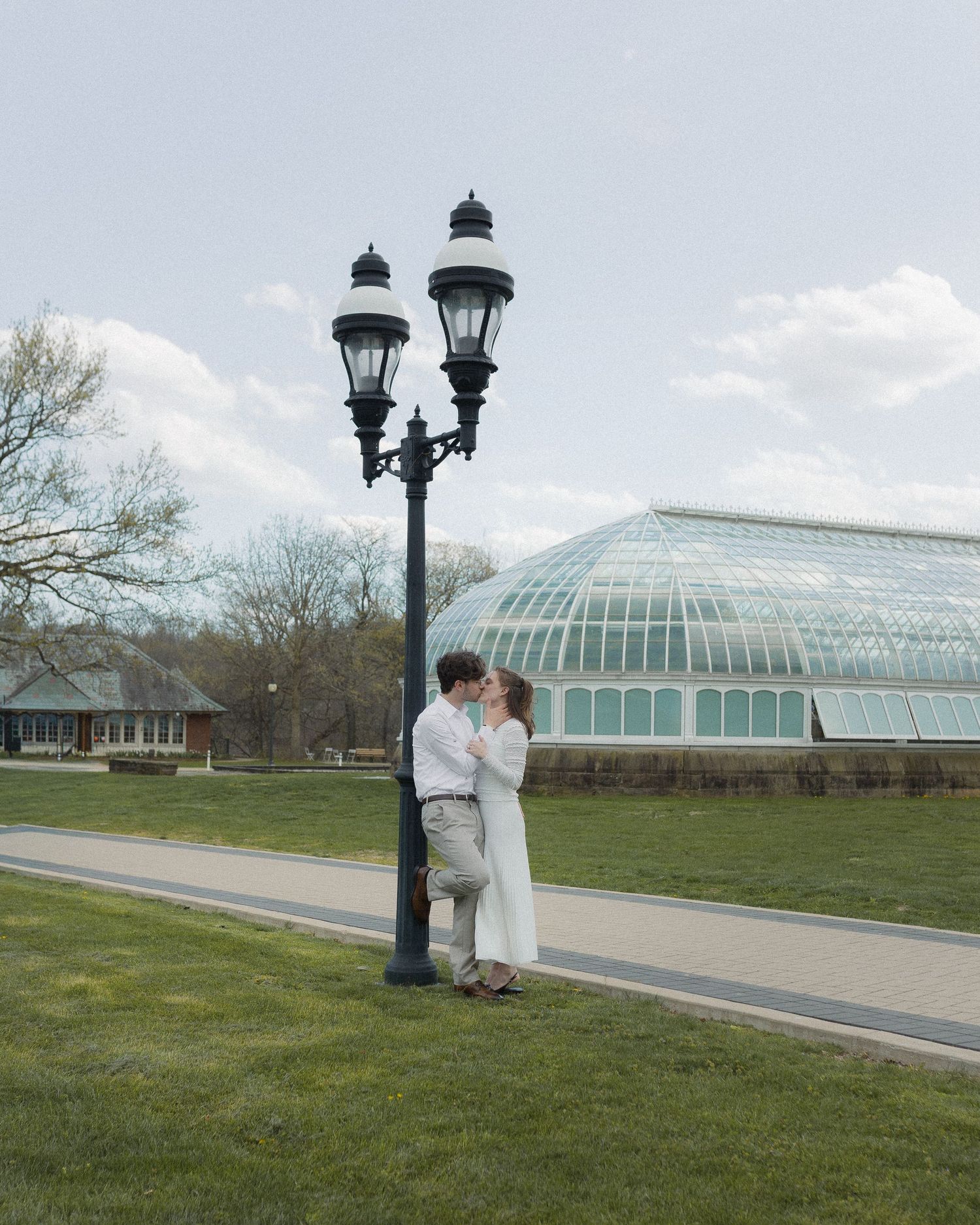 Couple kissing by a vintage lamp post with a glass greenhouse and green lawn in the background.