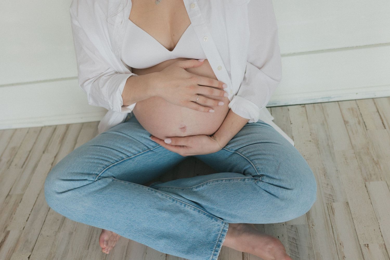 Pregnant woman in white top and jeans sits cross-legged on wooden floor holding her baby bump.