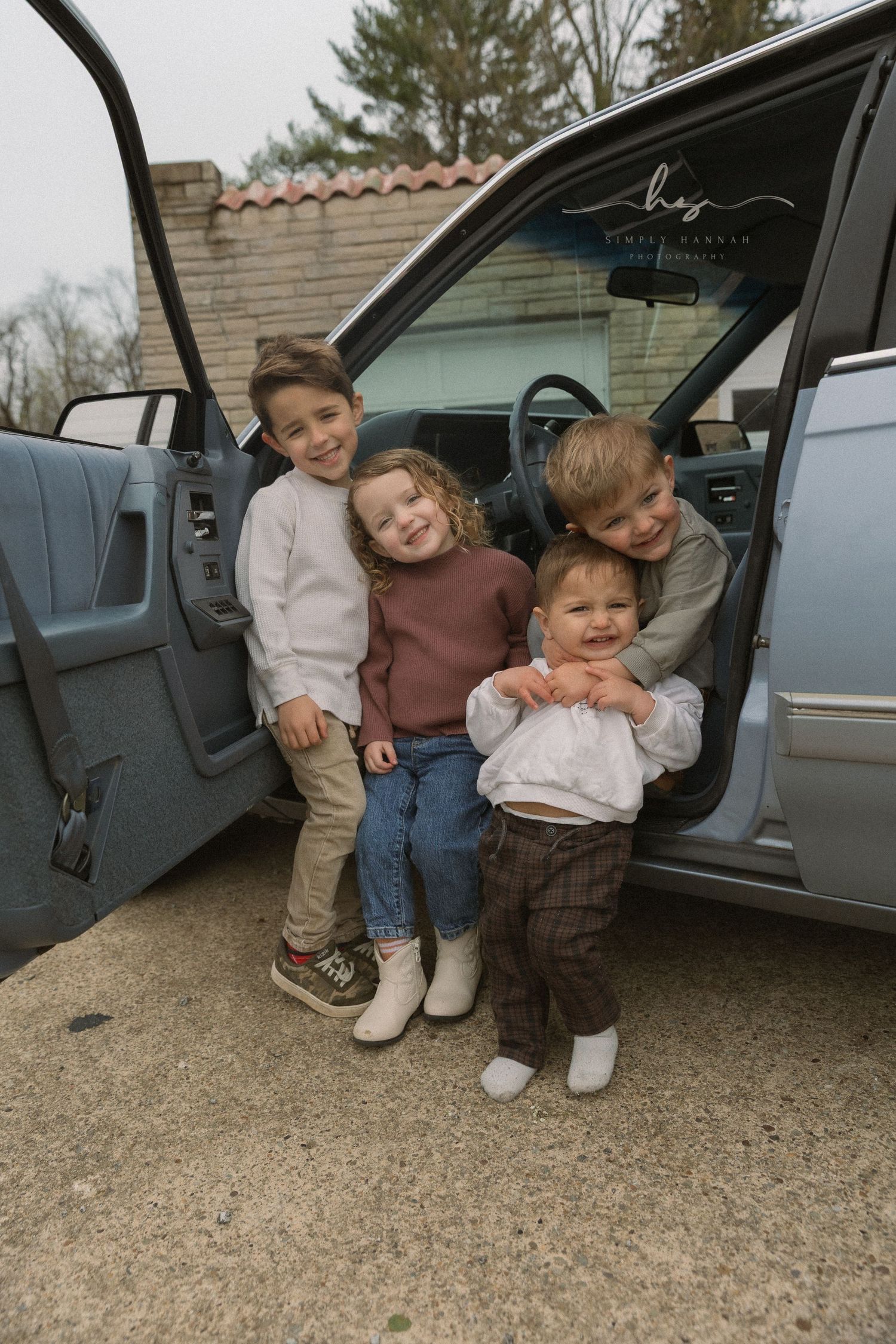 Candid lifestyle photo of four young siblings laughing, huddled by a vintage car, cozy family photography with neutral tones & real moments.
