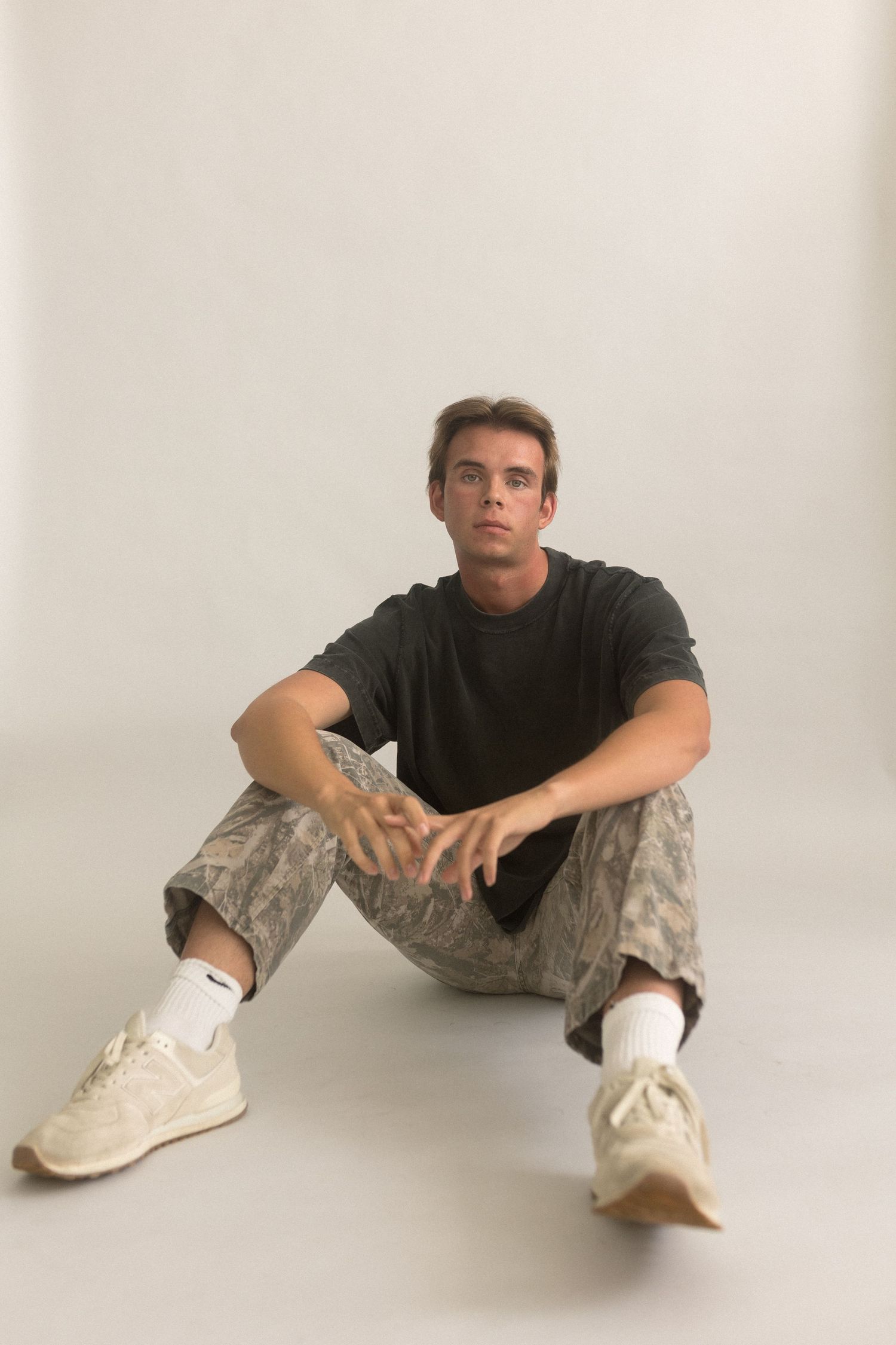 Person sits on floor wearing black t-shirt, camo pants and white sneakers in studio setting.