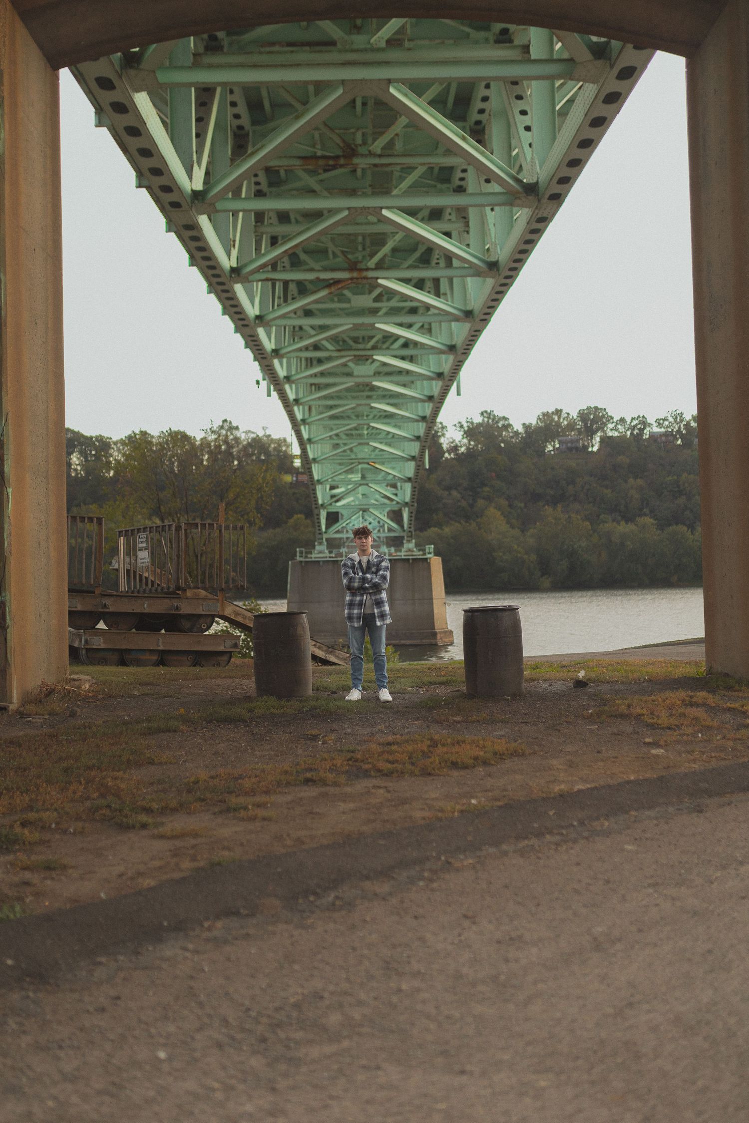 Person standing under geometric green bridge structure near river.