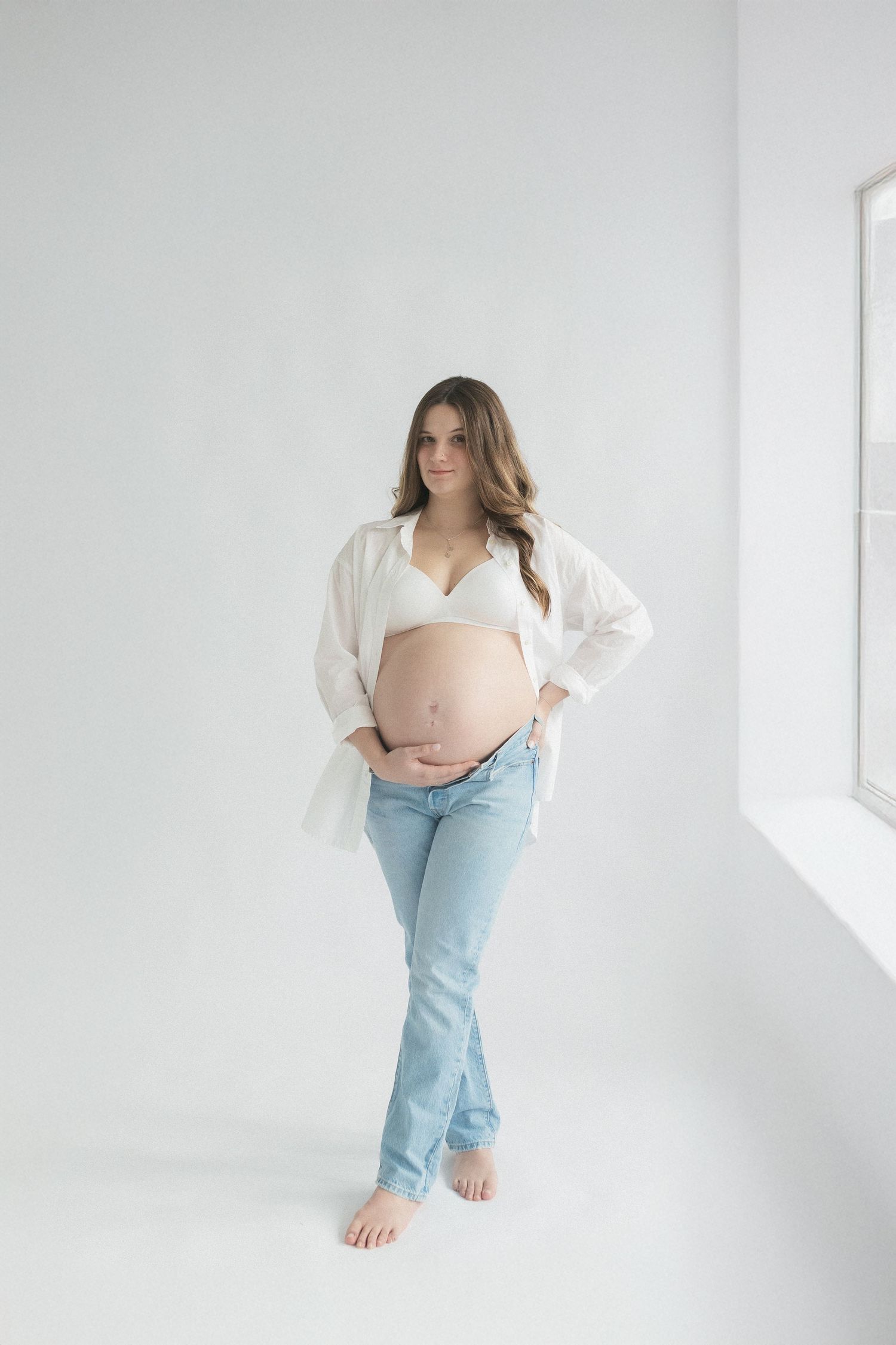 Expectant mother posing in casual white shirt and jeans in bright studio setting.