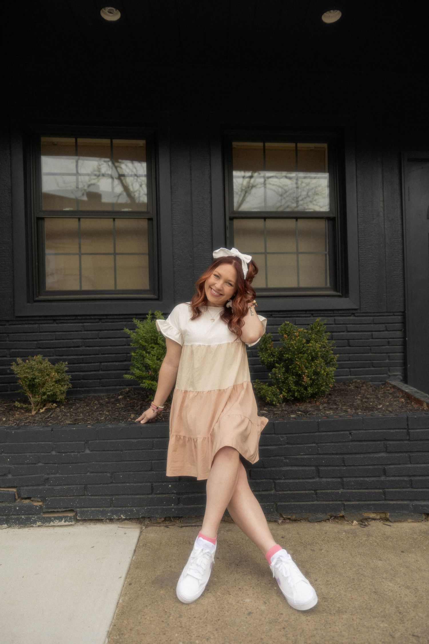Playful pose in tiered peach babydoll dress and white sneakers against black brick wall.