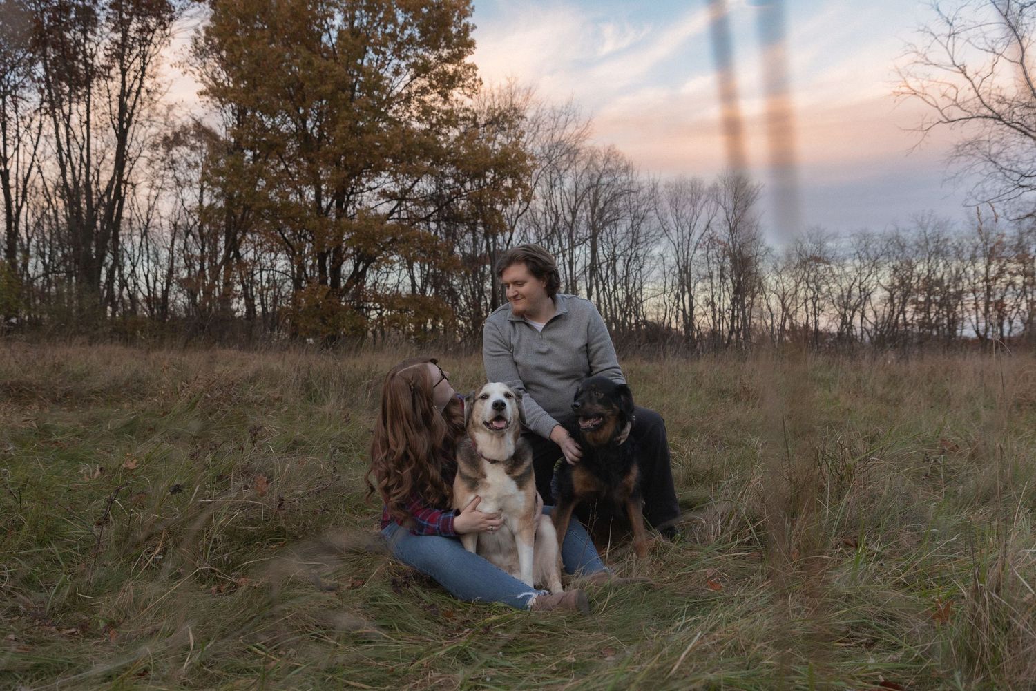Two people sitting with their dogs in a grassy field at dusk.