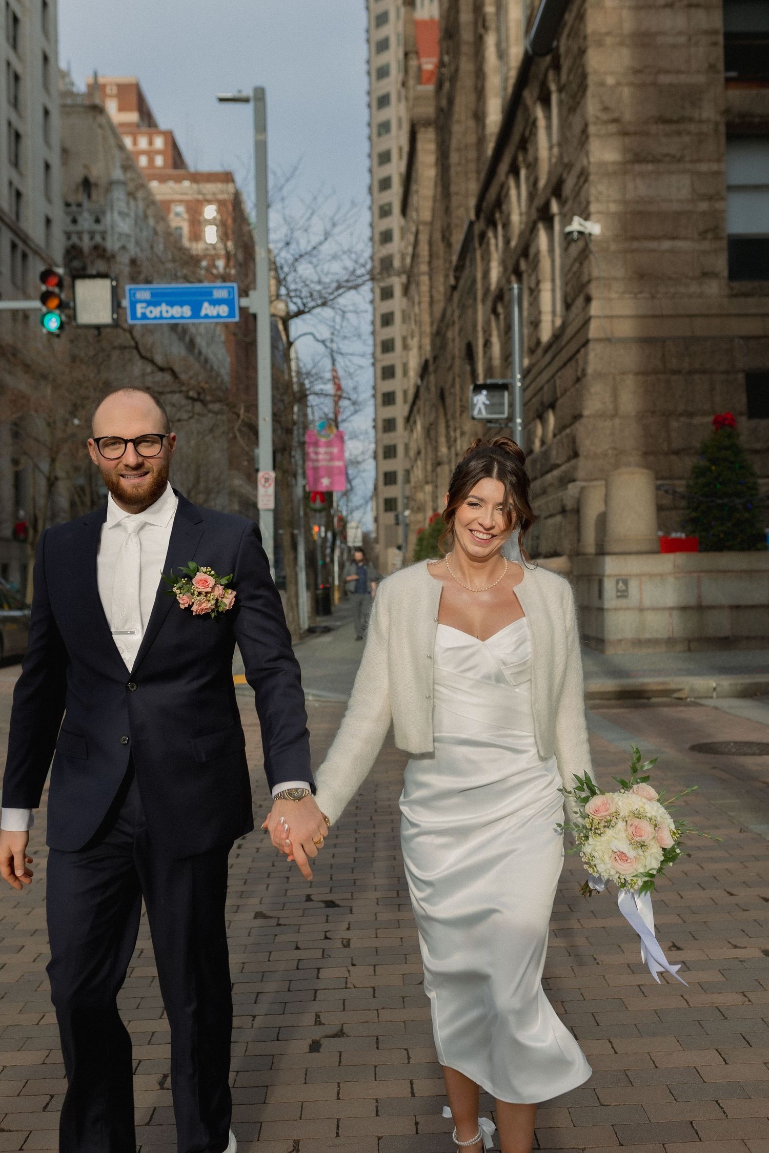 Newlyweds walking together on city sidewalk holding hands in elegant attire.
