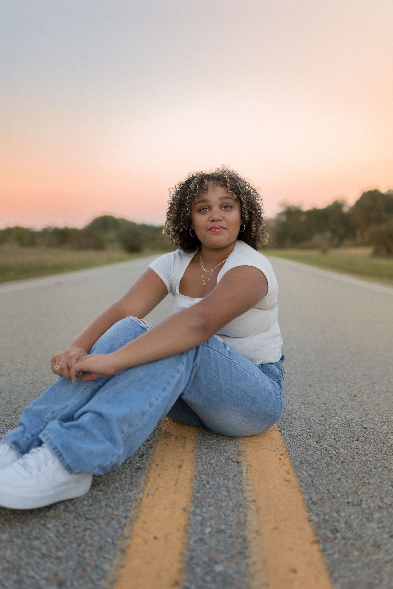 Person sitting on yellow road lines wearing white top and blue jeans at sunset.