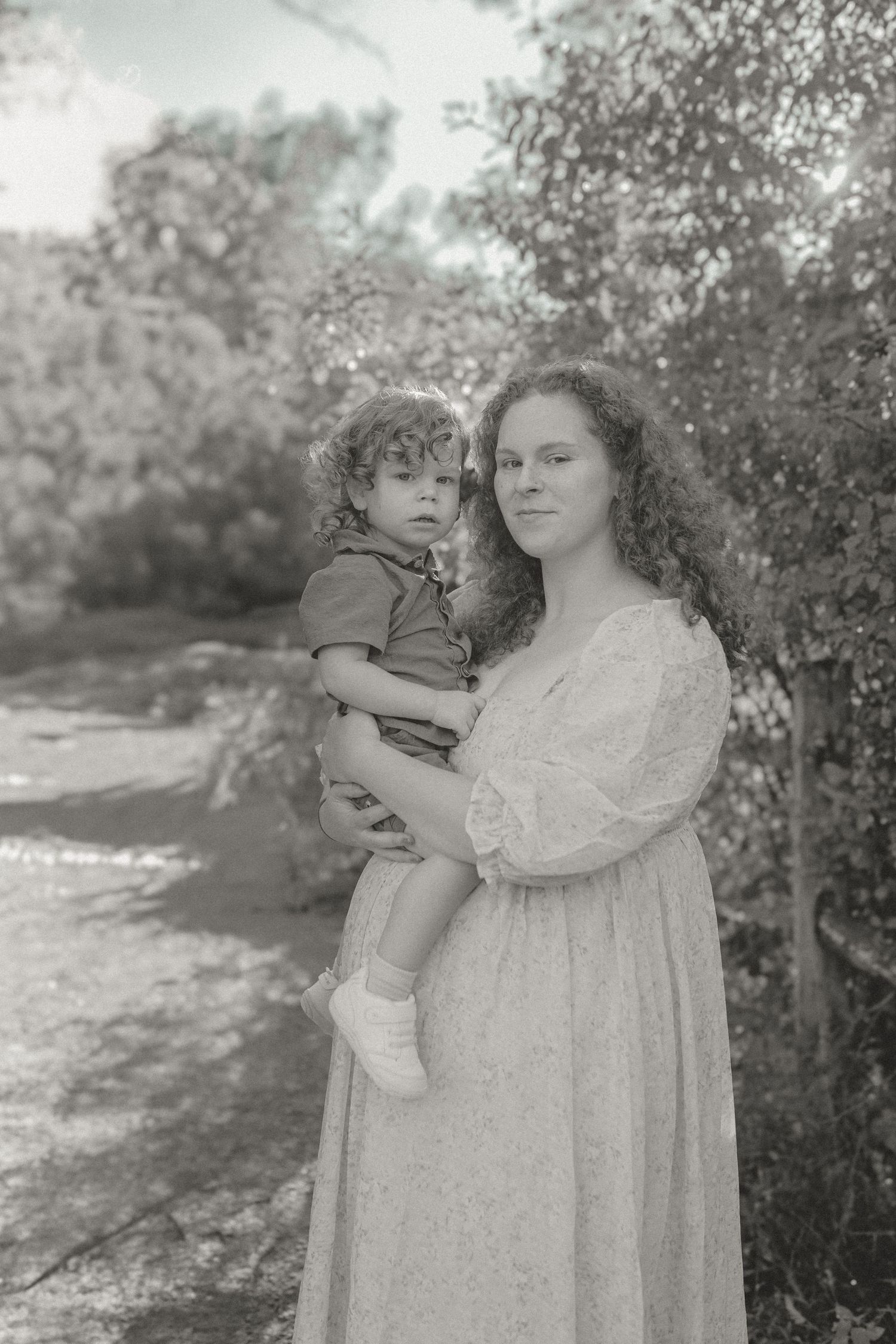 Black and white portrait of a woman in a white dress holding a toddler outdoors.