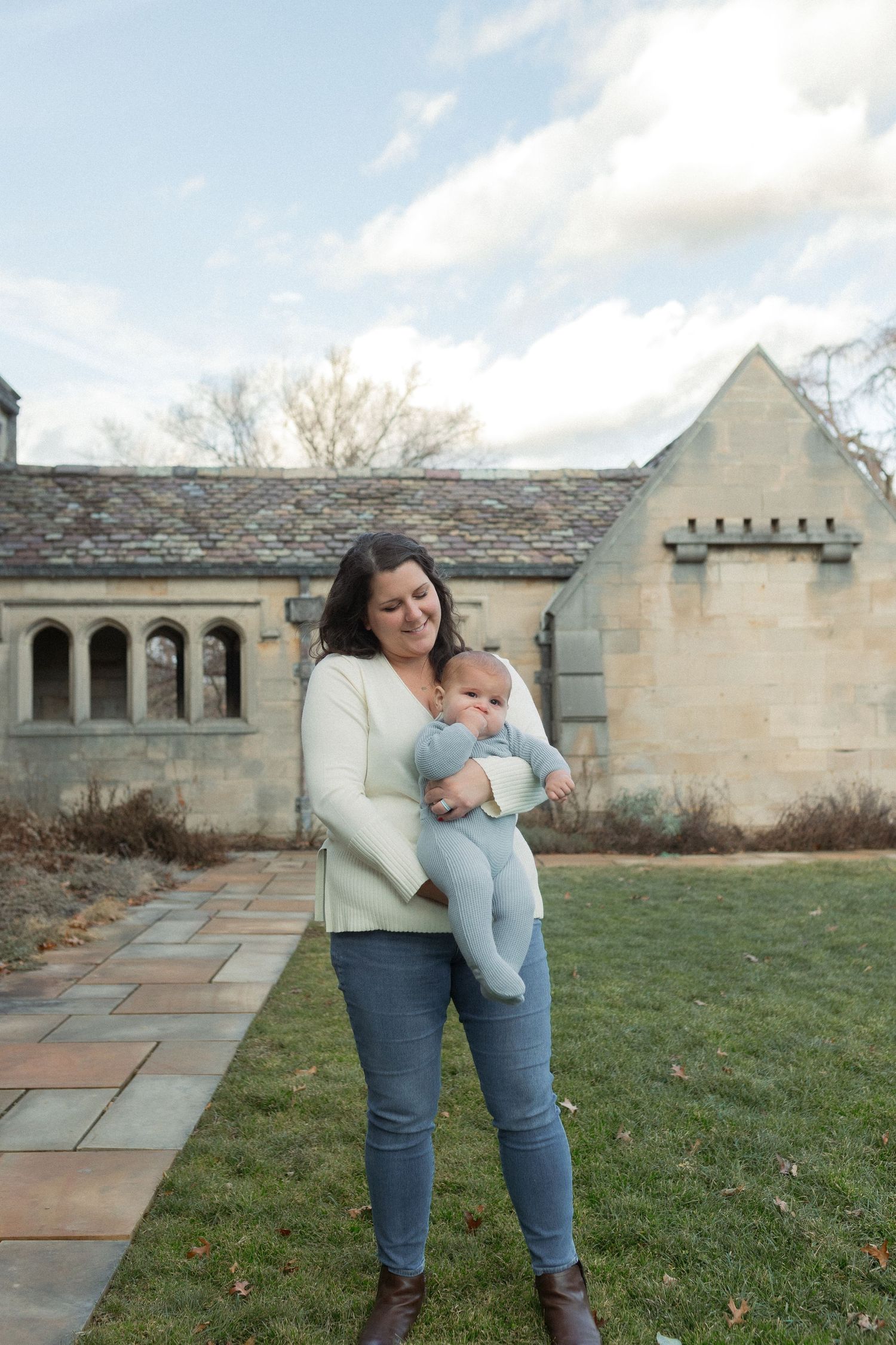 Woman in white sweater holding an infant outside a stone building with arched columns on a cloudy day.