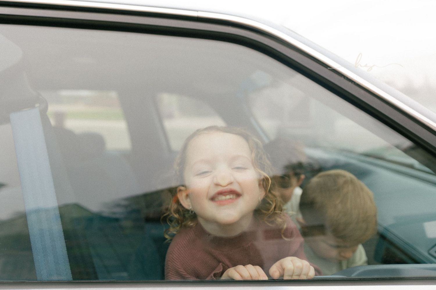 Toddler grinning through a car window, candid childhood photography with genuine smiles and natural light storytelling.