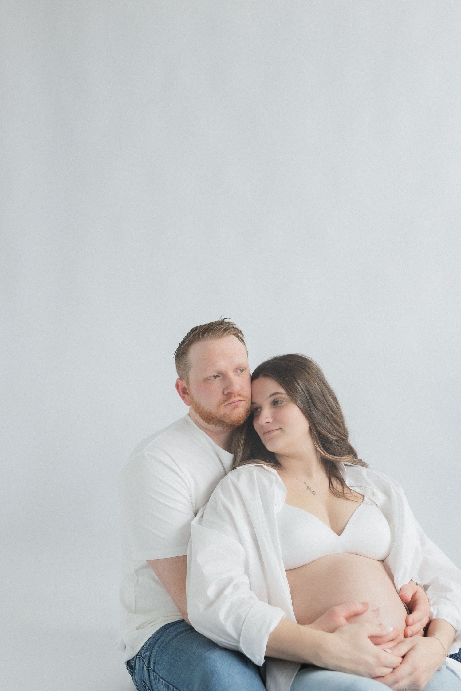 Intimate maternity portrait with couple embracing against minimalist white backdrop.