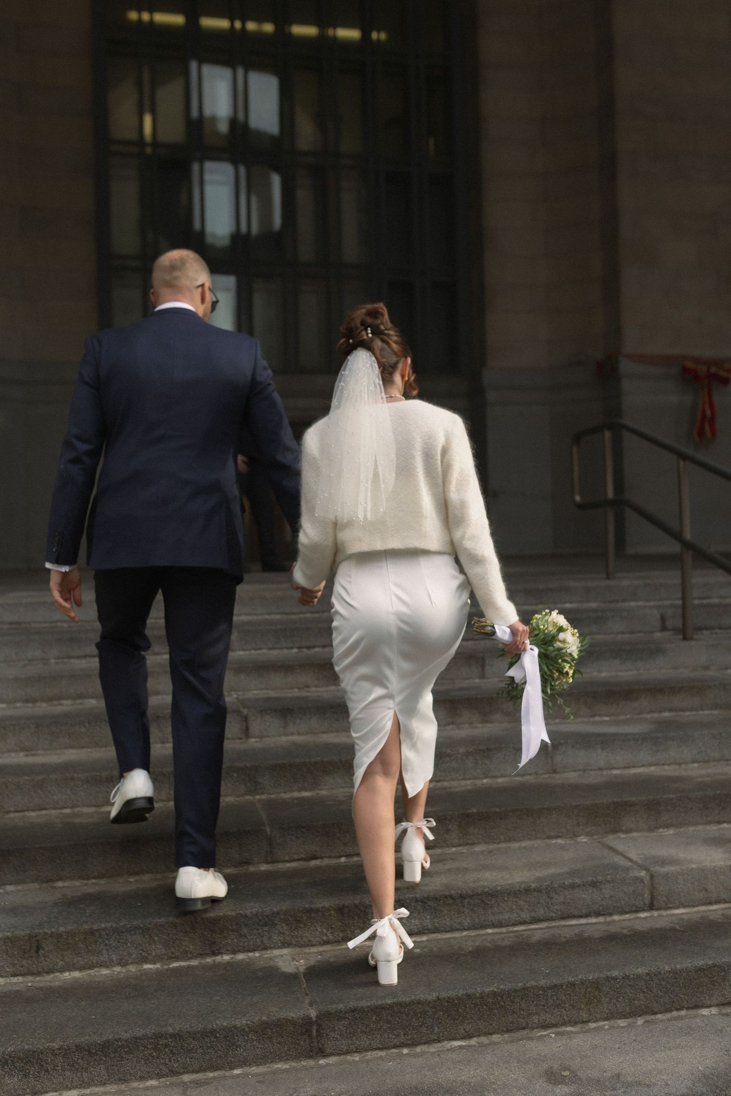Couple in wedding attire walking up stone steps together.
