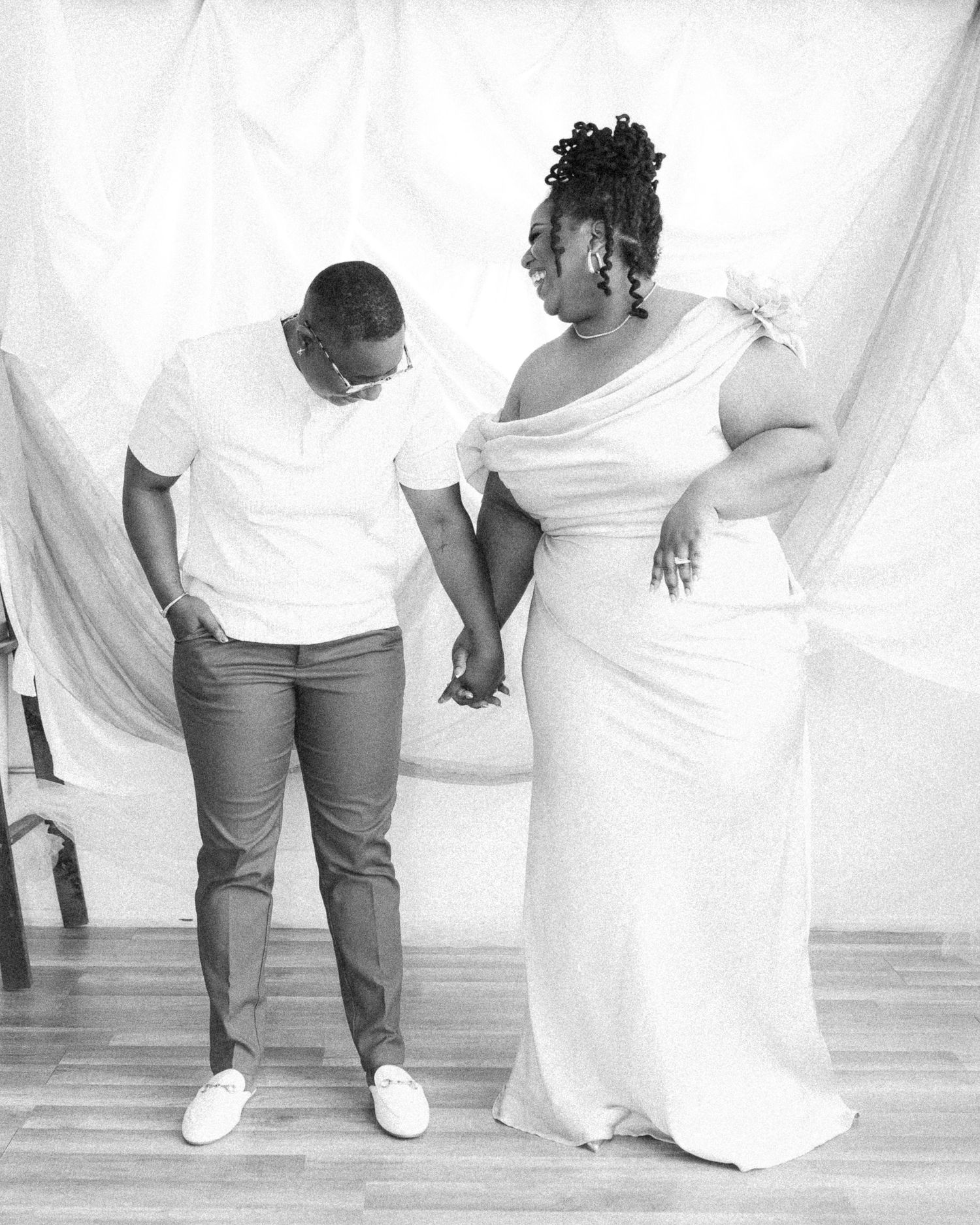 A couple in white outfits share a tender moment while holding hands against a light backdrop during a photoshoot.