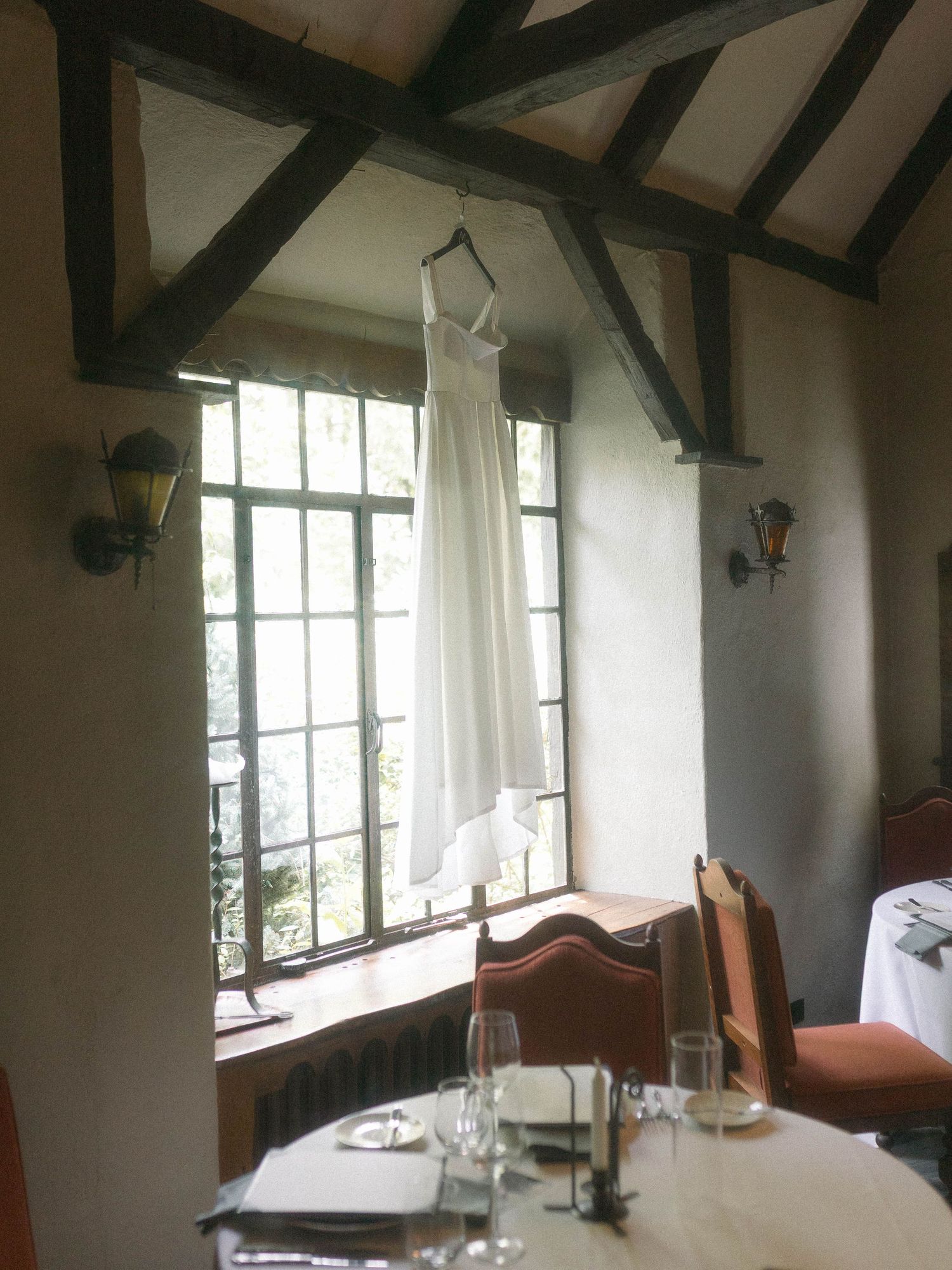 A white wedding dress hangs by a window in a rustic room with exposed wooden beams and an elegant table setting.