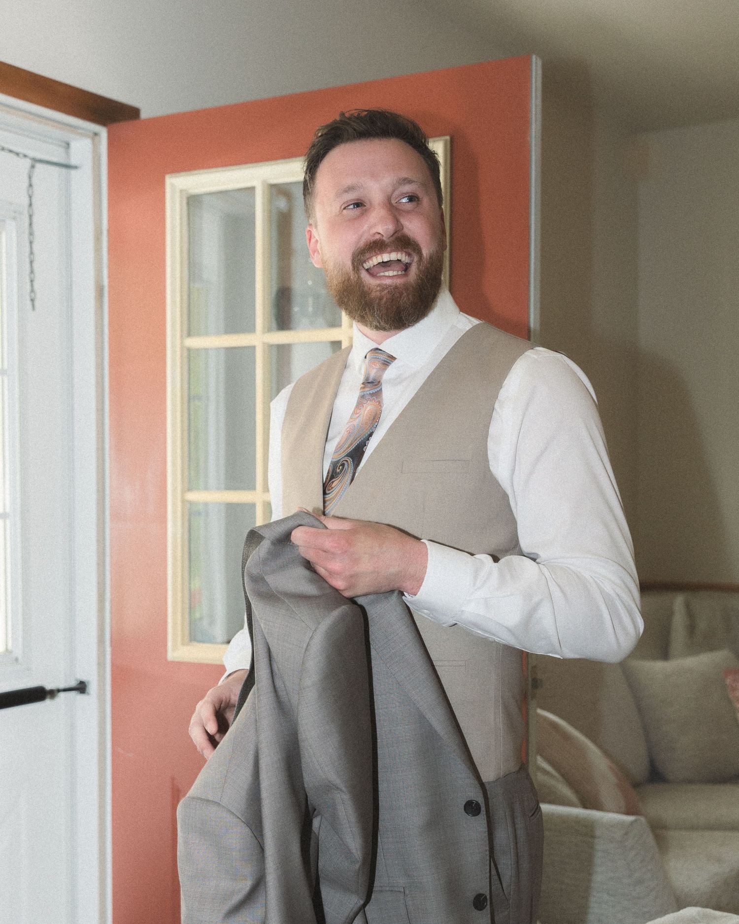 A smiling person wearing a tan vest and white dress shirt buttons up formal attire in an interior setting with coral walls.