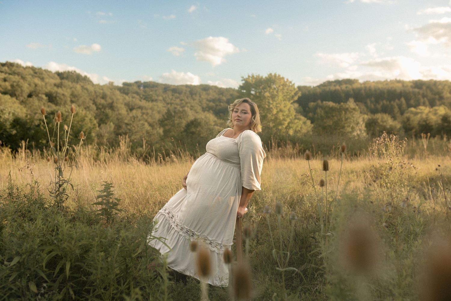 Maternity photo of woman in white dress standing in golden sunset field.